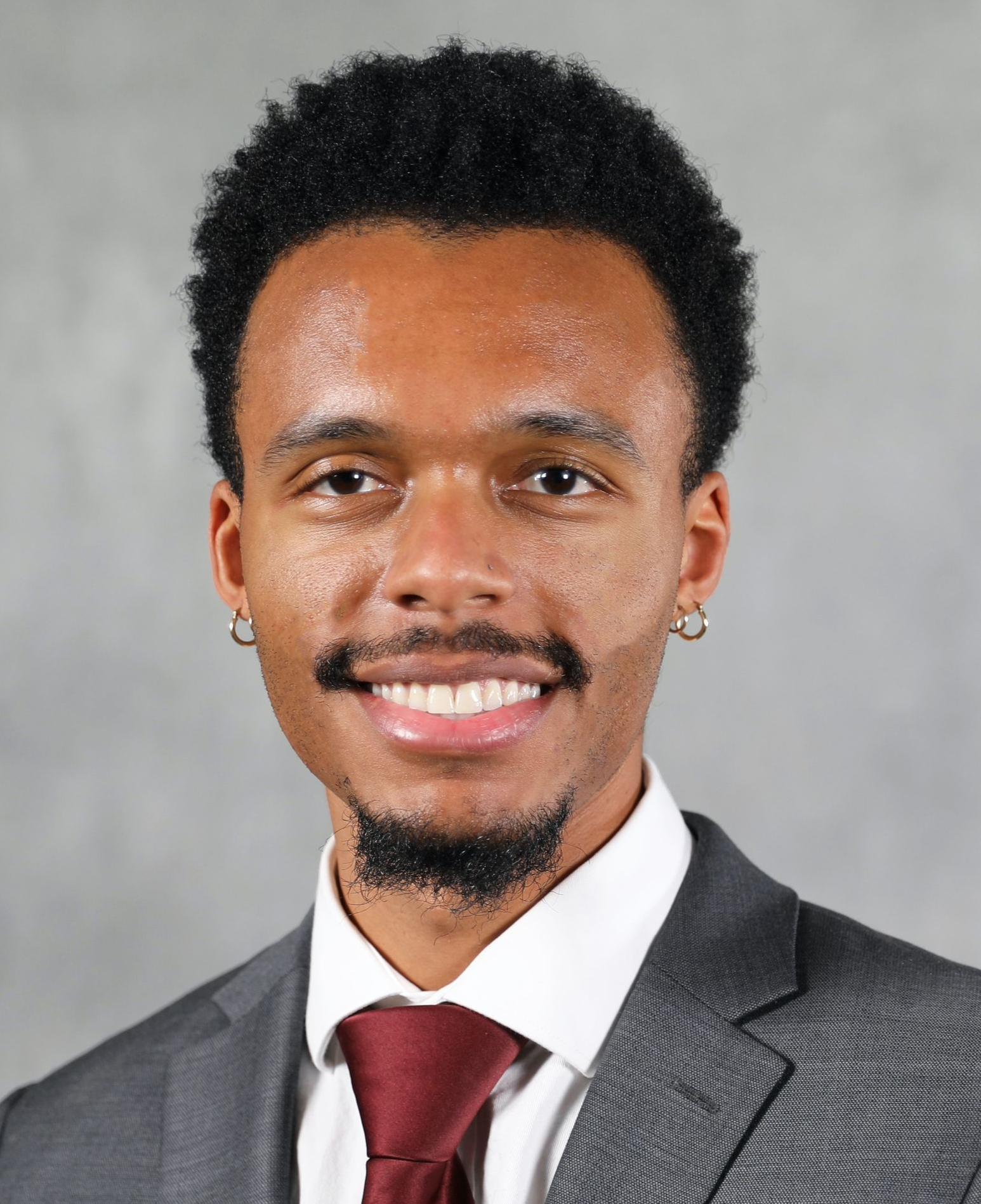 A smiling individual wearing a dark suit, white shirt, and maroon tie against a neutral grey background.