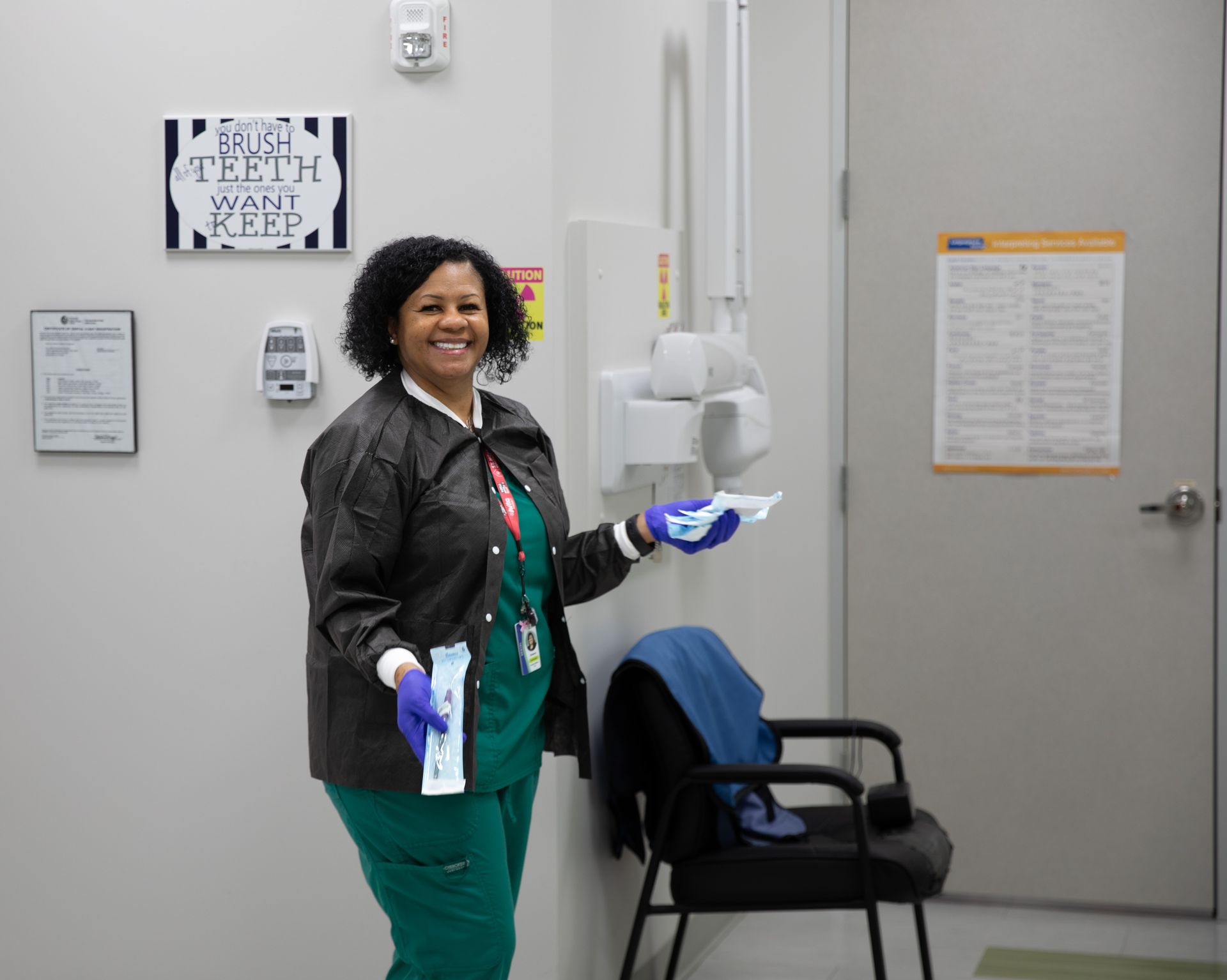 A smiling healthcare worker in green scrubs and a black jacket holds medical supplies in a dental office.