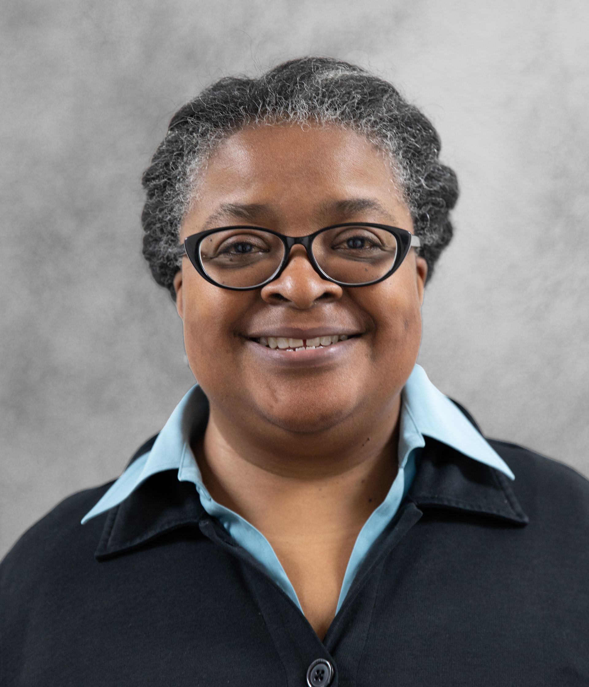 A person with short, gray-streaked hair wears glasses, a light blue collared shirt, and a dark cardigan against a gray wall.