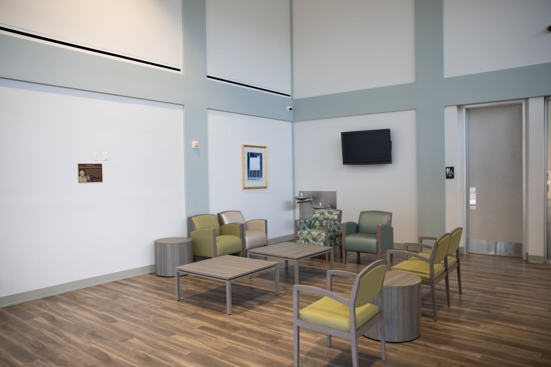 A bright waiting room with pale green and tan chairs, two small square tables, wood-look flooring, and a wall-mounted TV.