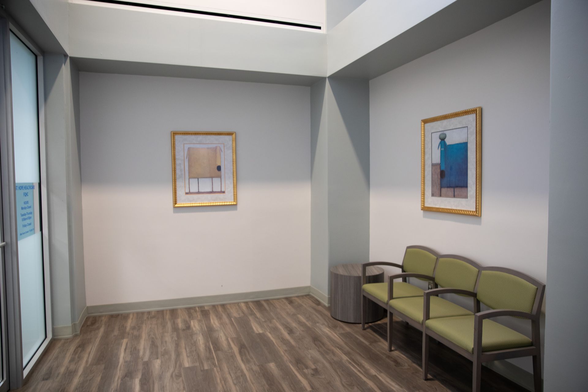 A waiting area with two framed artworks on light walls, wood-look flooring, and a green three-seat chair.