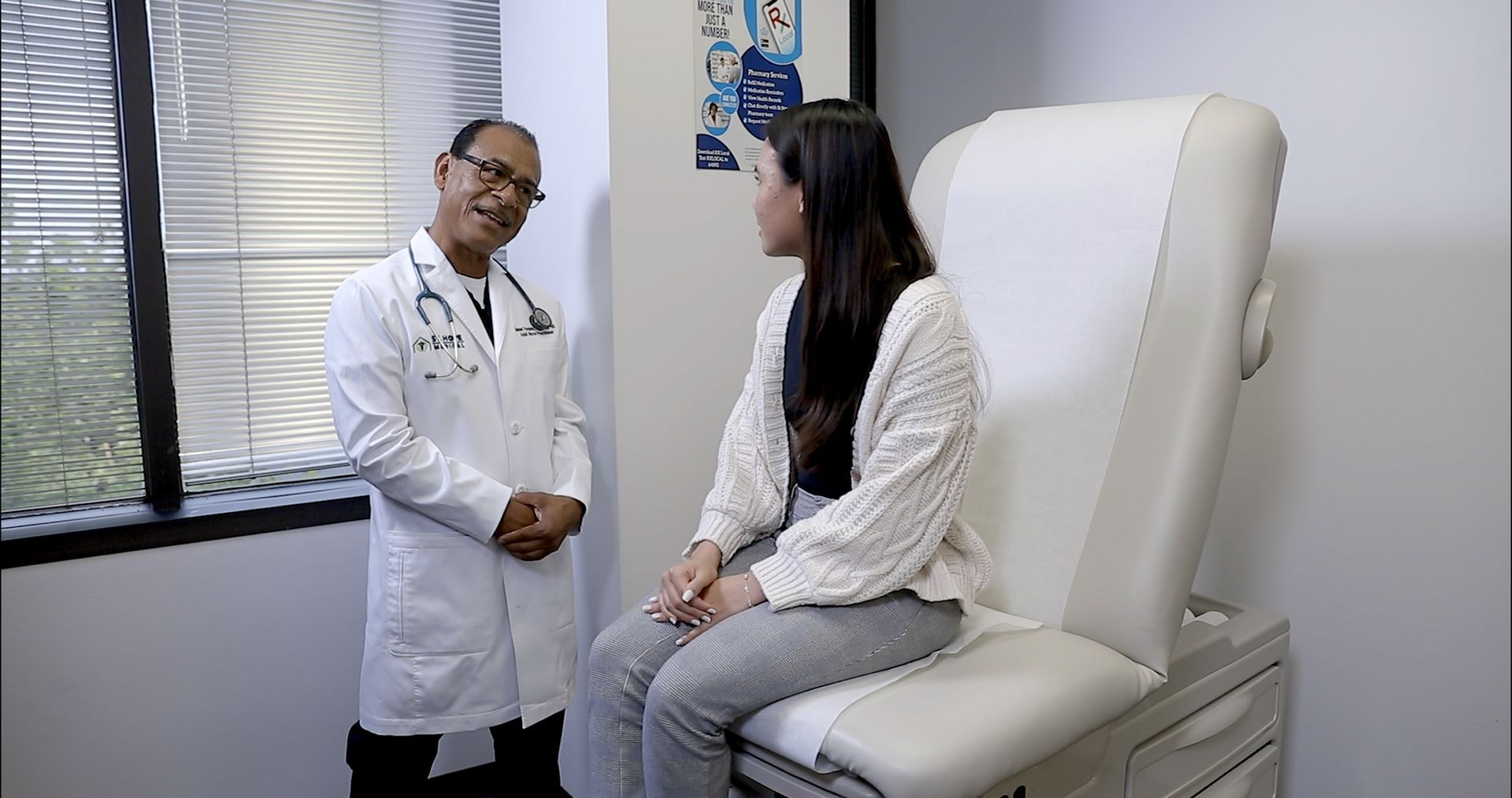 A doctor in a white coat talks to a patient seated on an exam table in a clinical office.