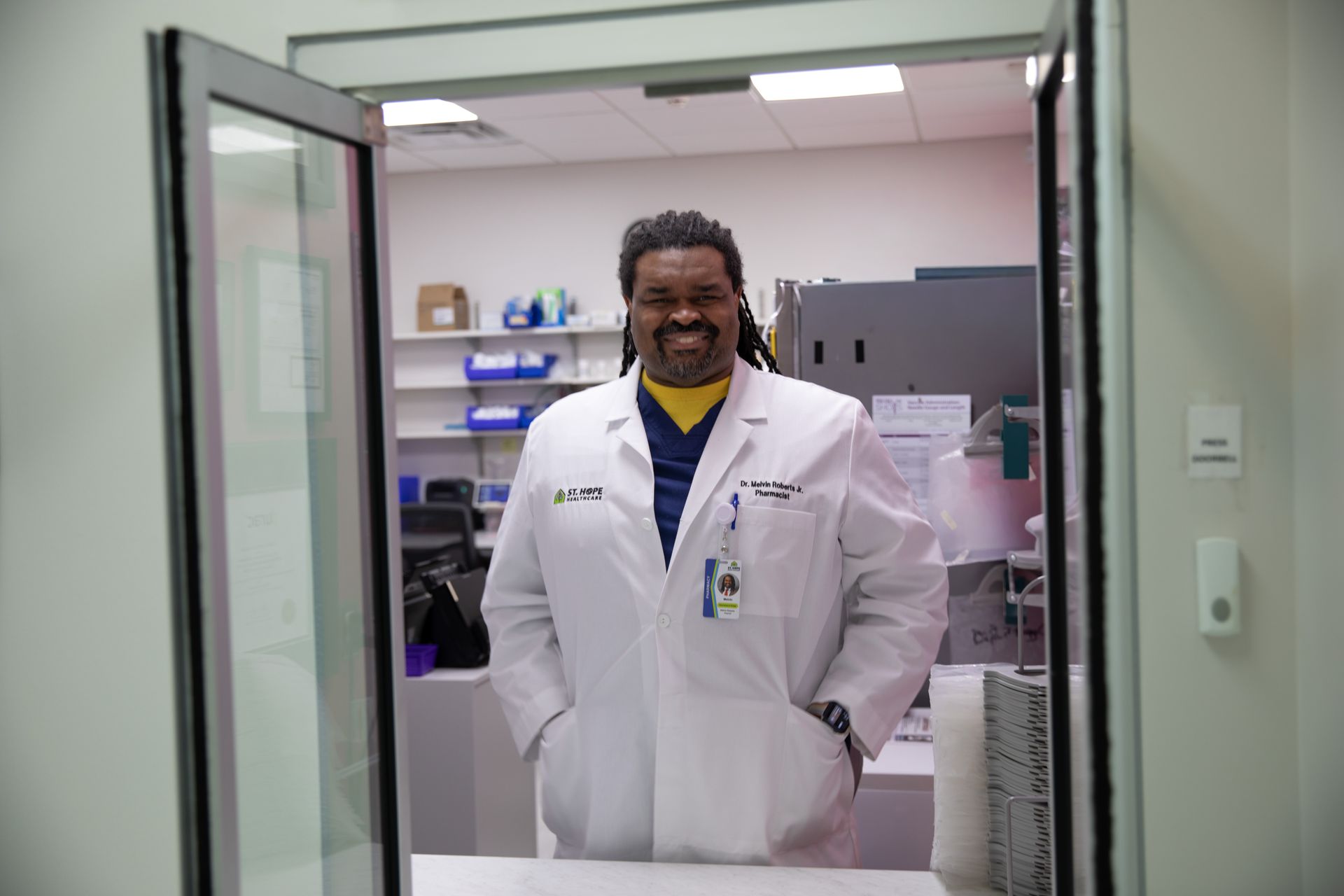 A smiling person in a white lab coat stands with hands in pockets in a pharmacy or medical workspace.