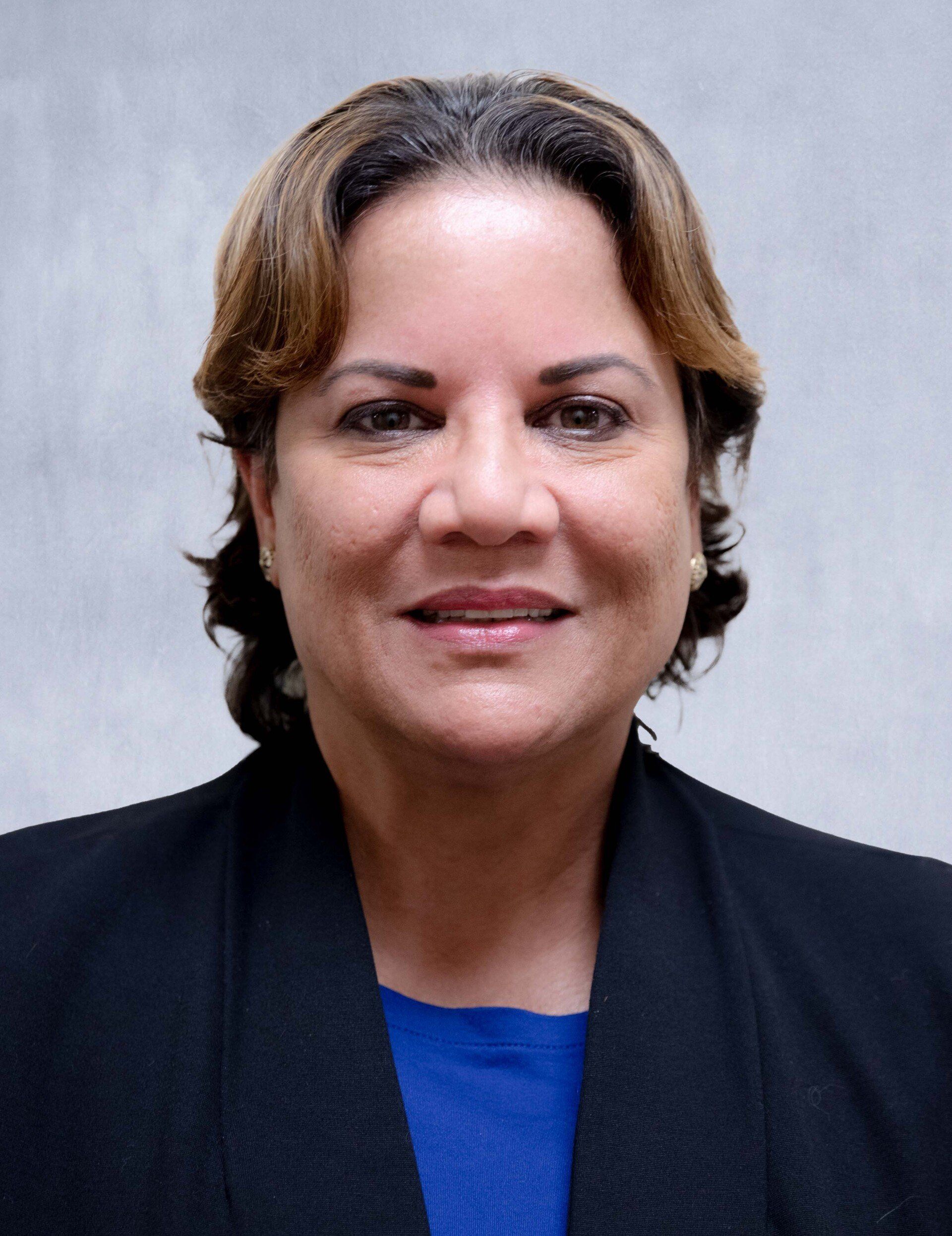 A professional headshot of a person with shoulder-length brown hair, wearing a blue shirt under a black blazer.