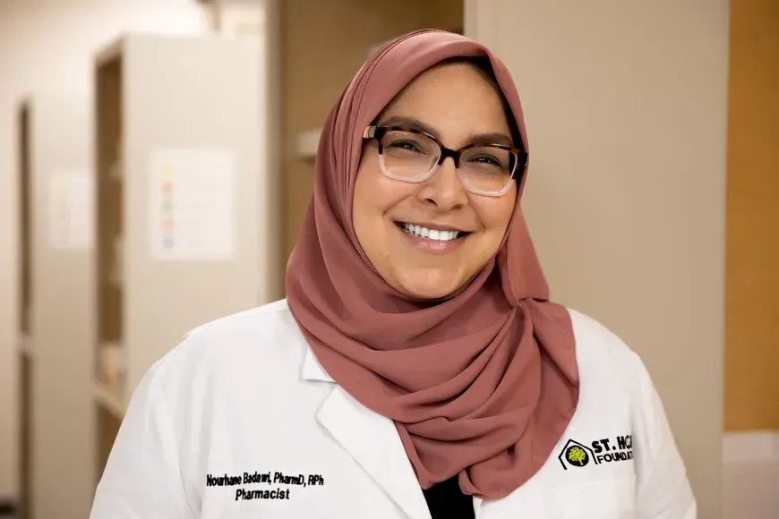 Smiling pharmacist wearing a pink hijab, glasses, and a white lab coat in an office setting.