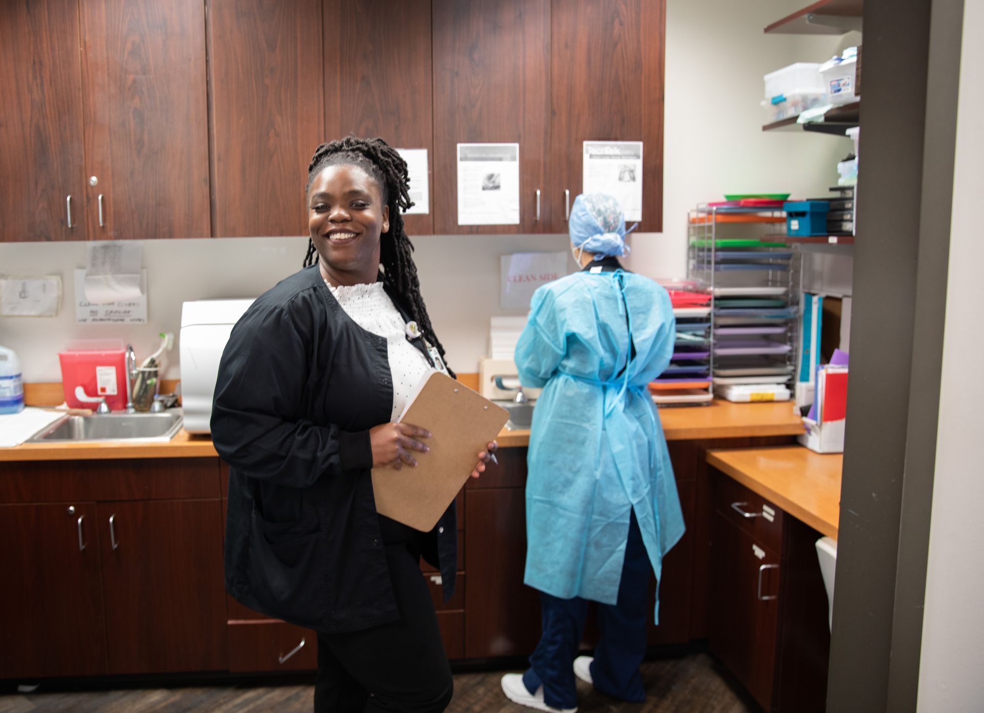 A staff member in a black uniform holds a clipboard in a medical office, while a colleague in blue scrubs works nearby.