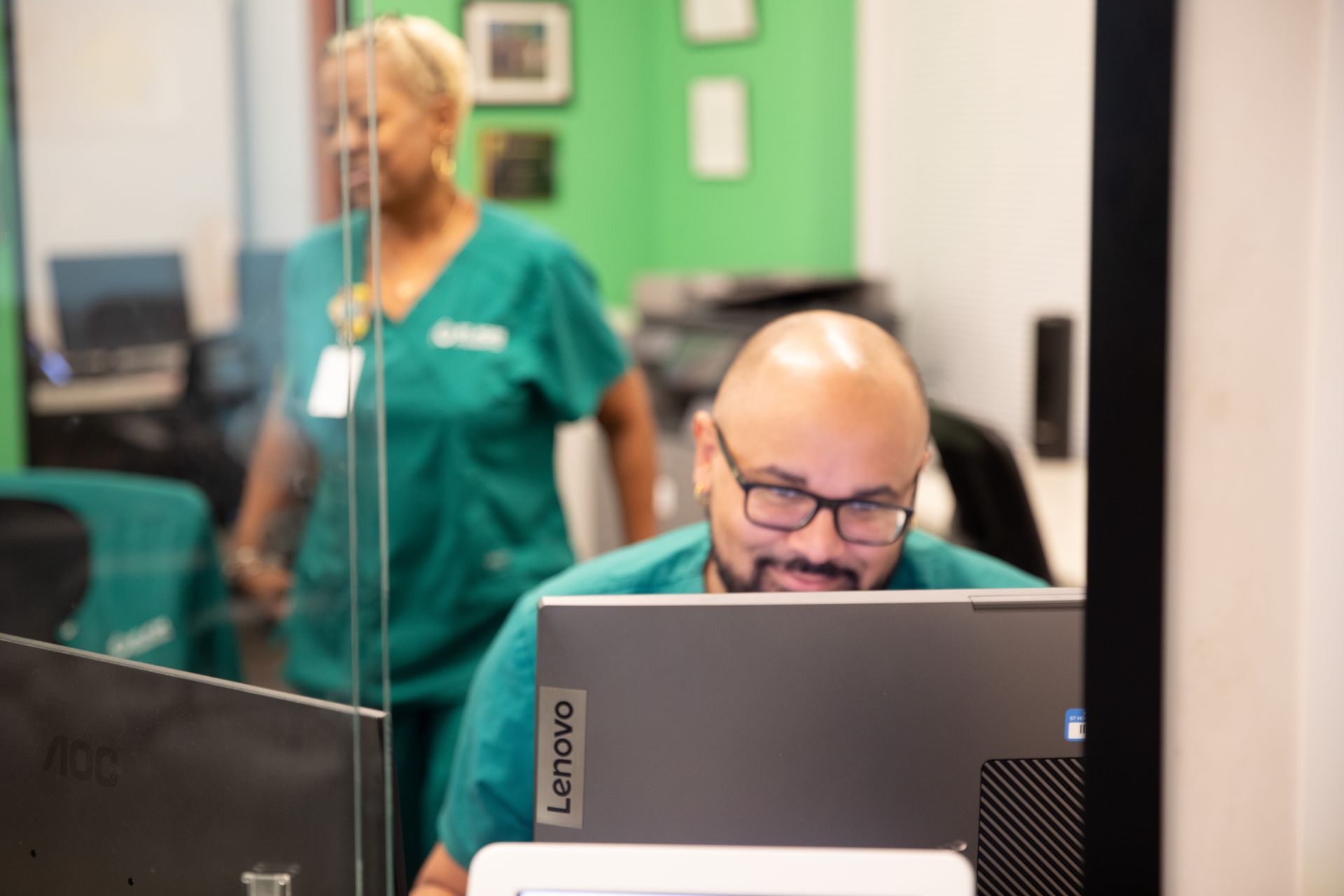A staff member in teal scrubs works at a computer in an office, while a colleague walks in the background.