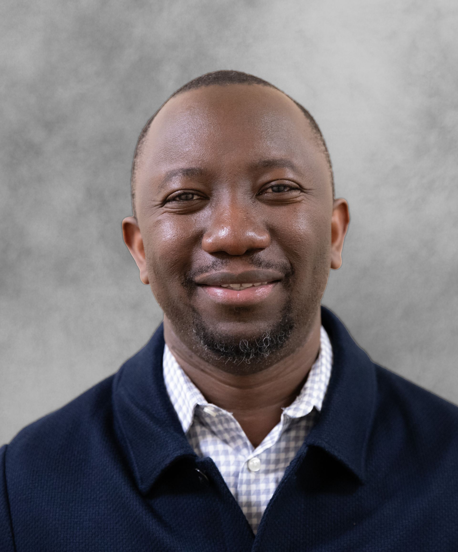 A smiling individual wearing a collared, checkered shirt under a dark jacket against a plain grey background.