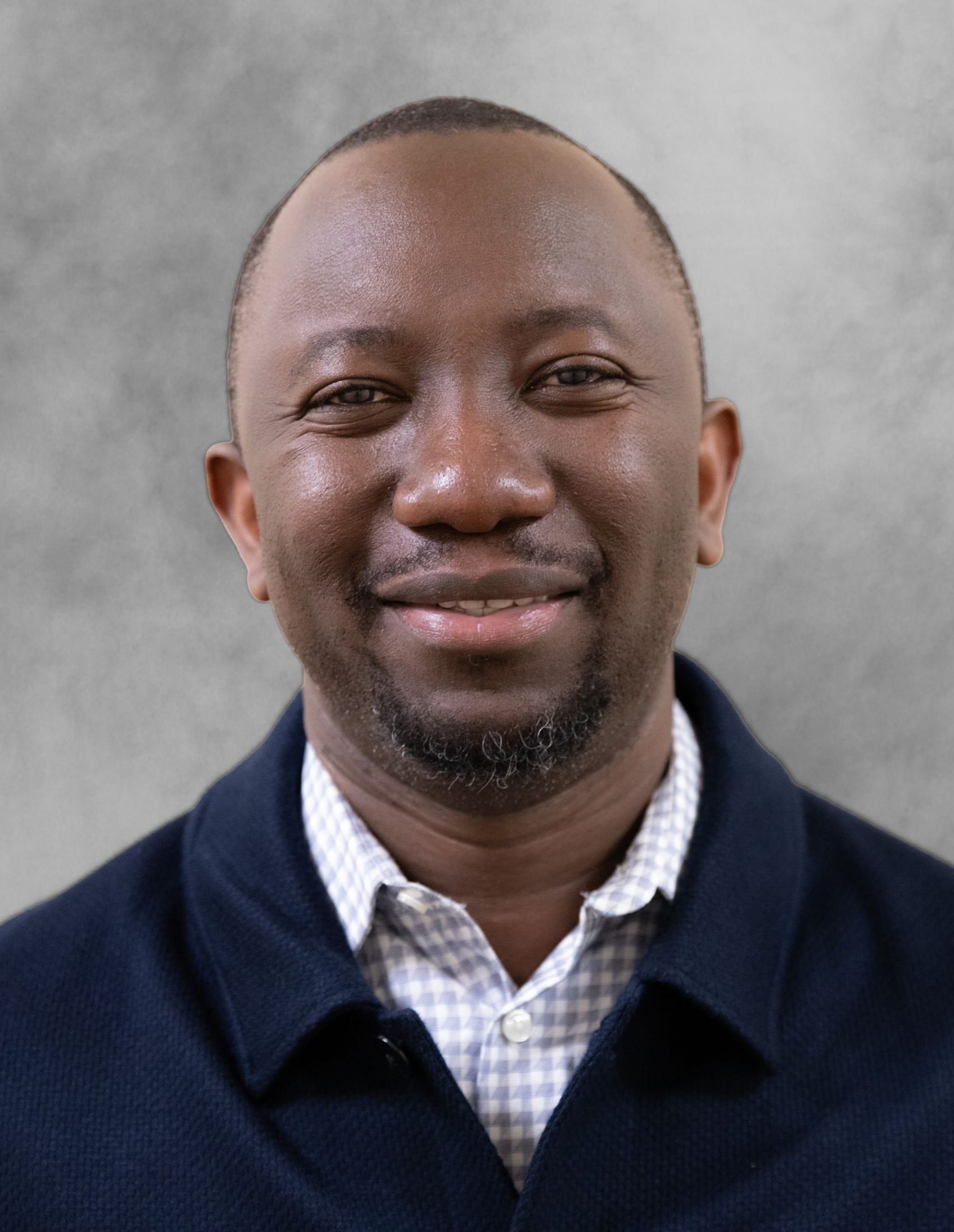 A man with a short beard smiles at the camera, wearing a blue sweater over a light, patterned collared shirt.