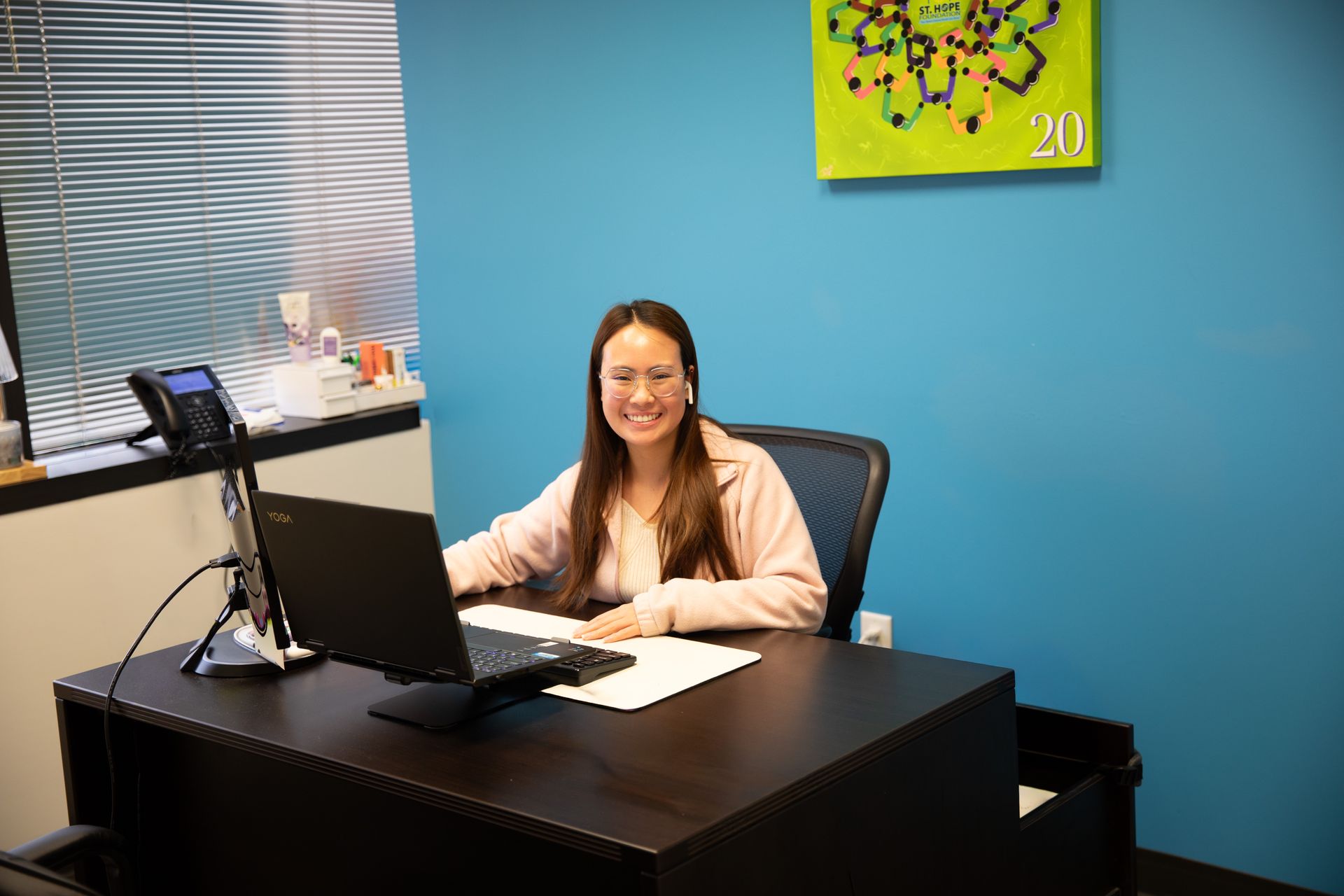 A smiling person wearing a headset sits at a desk with a laptop in an office with blue walls and a window with blinds.