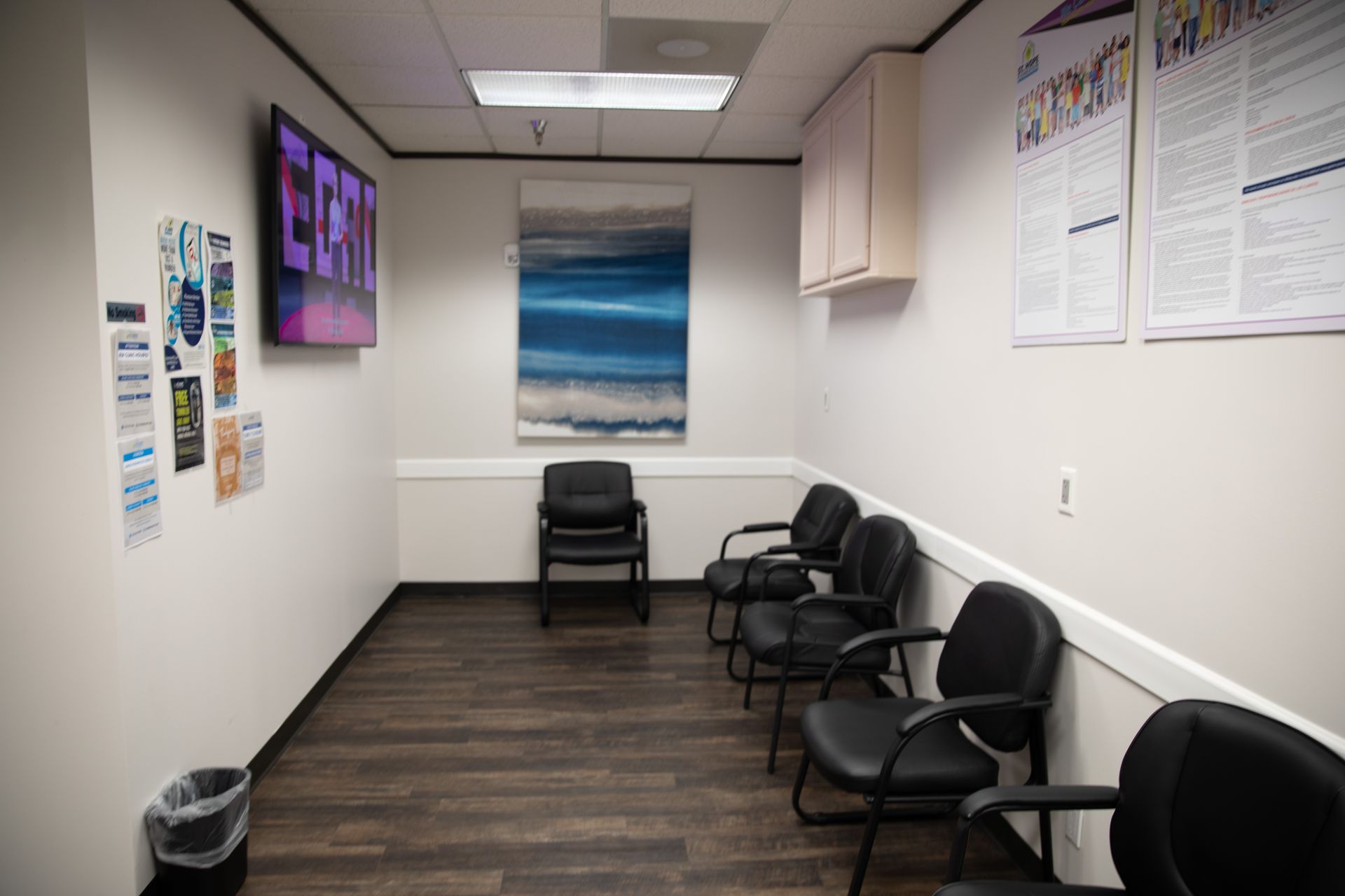 A waiting room with five black chairs, a wall-mounted television, an abstract blue painting, and posters on the wall.