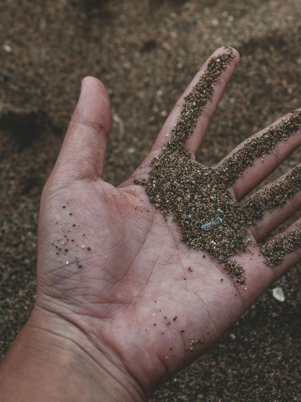A person's open hand holding coarse, dark sand against a blurred, sandy background.