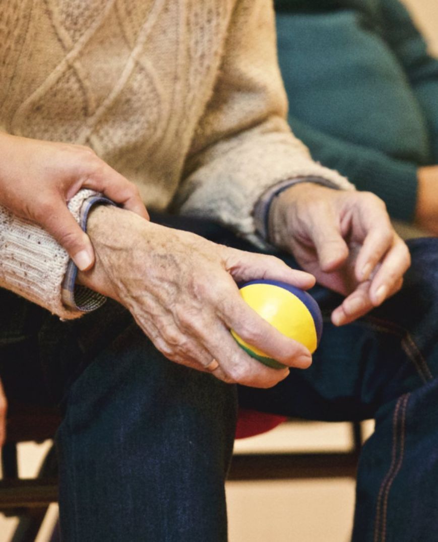 A person’s hand supports the wrist of another person who is holding a small, yellow and blue stress ball.