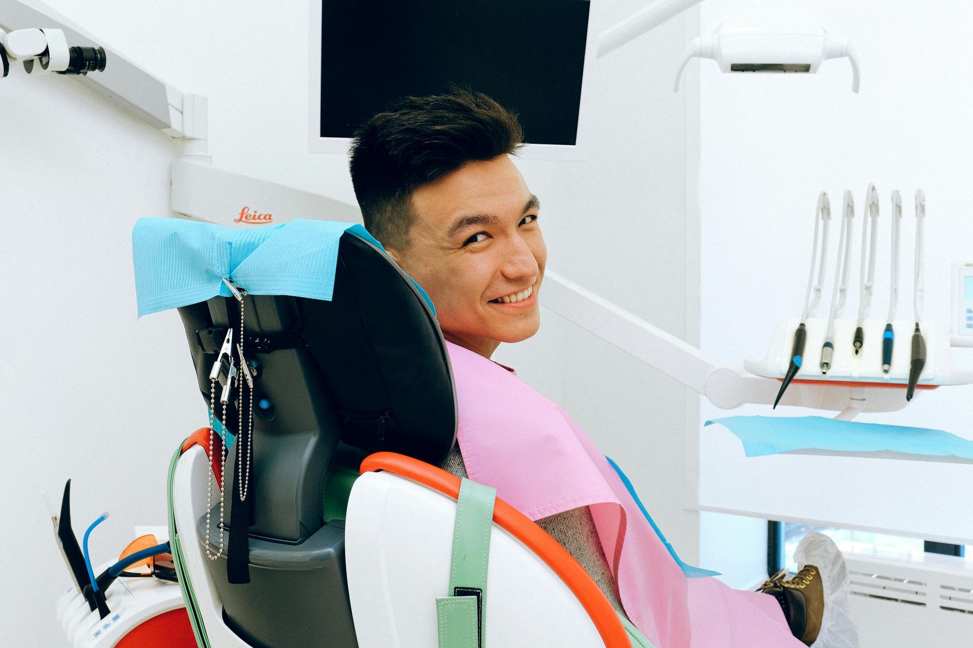 A person sits in a dentist's chair, wearing a pink bib and smiling at the camera in a brightly lit dental office.