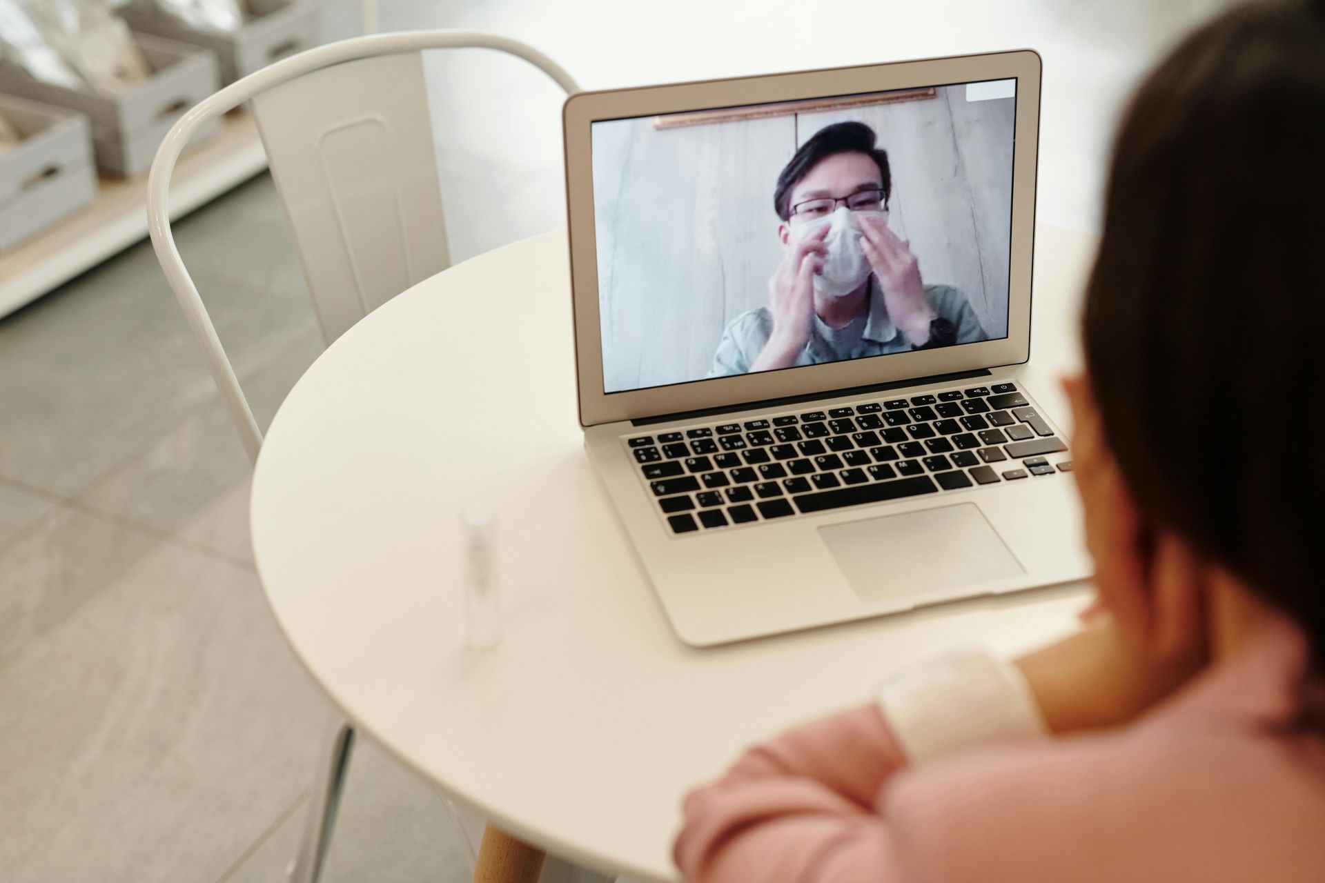 A person sits at a table and uses a laptop to video call someone who is adjusting a face mask.