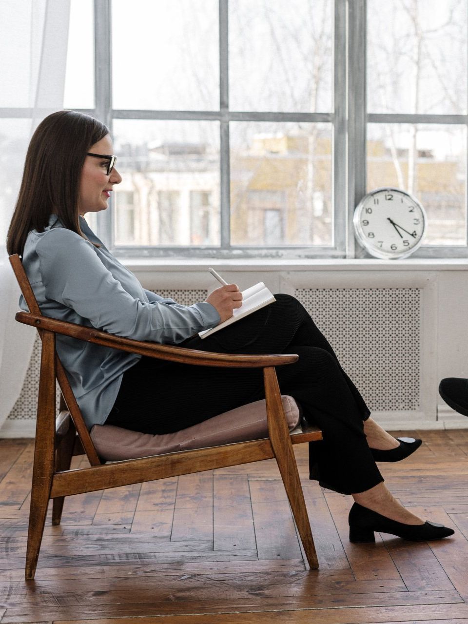 A person in professional attire sits in a wooden chair, holding a notebook and pen, facing an unseen person.
