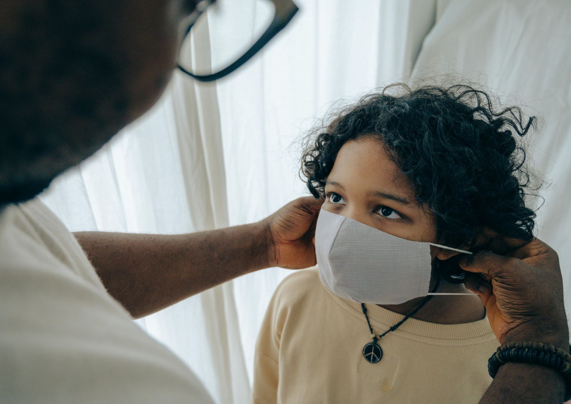 An adult’s hands adjust a light-gray fabric face mask on a person with curly hair wearing a pale yellow shirt.
