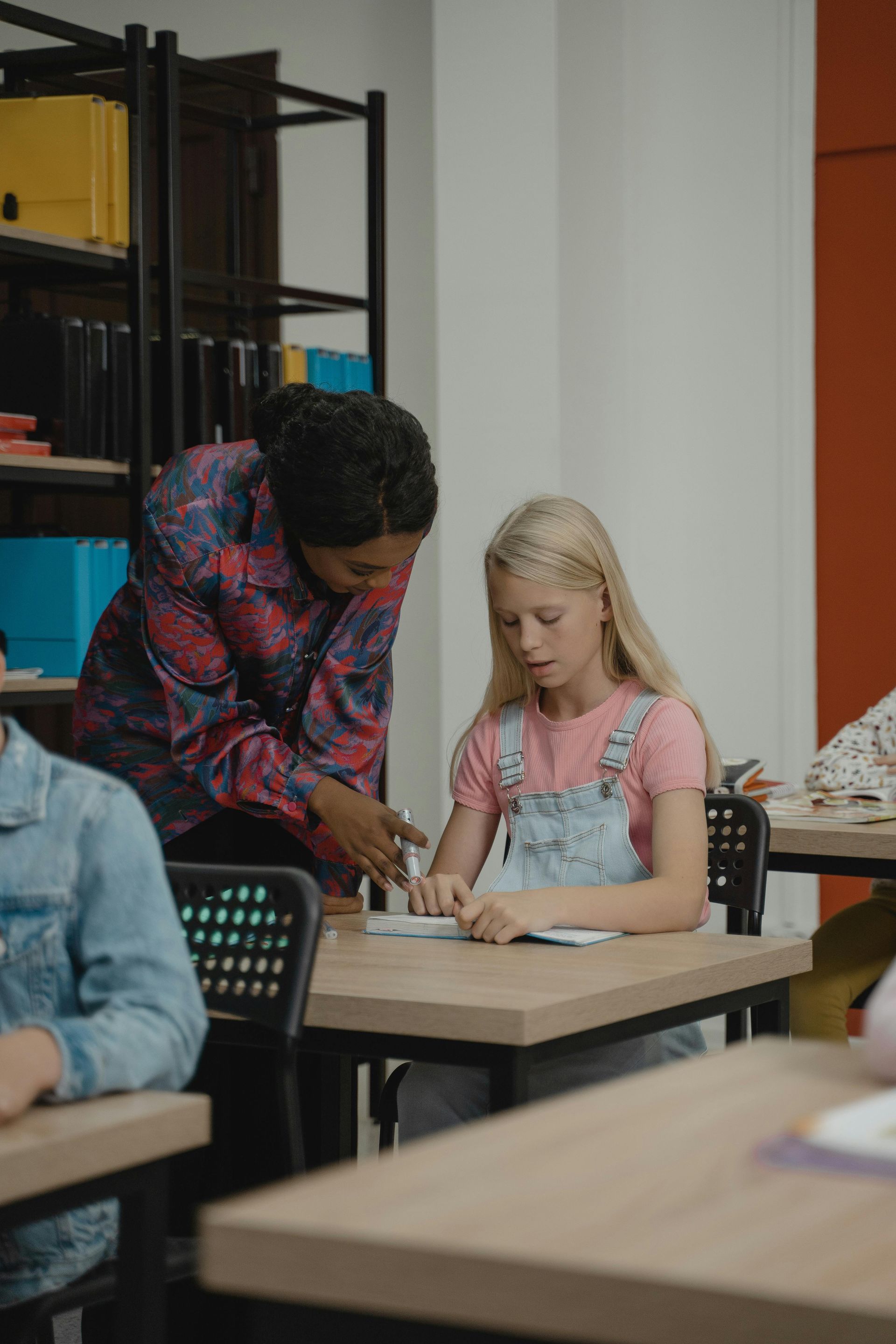 A teacher assists a student with schoolwork at a desk in a classroom.