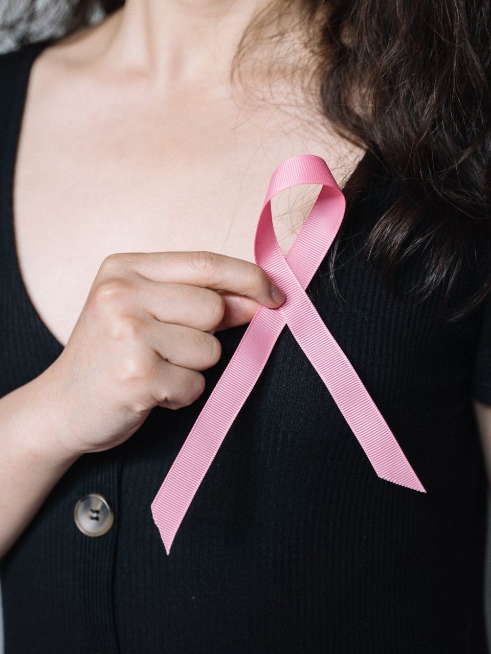 A person in a black shirt holds a pink ribbon against their chest, a symbol of breast cancer awareness.
