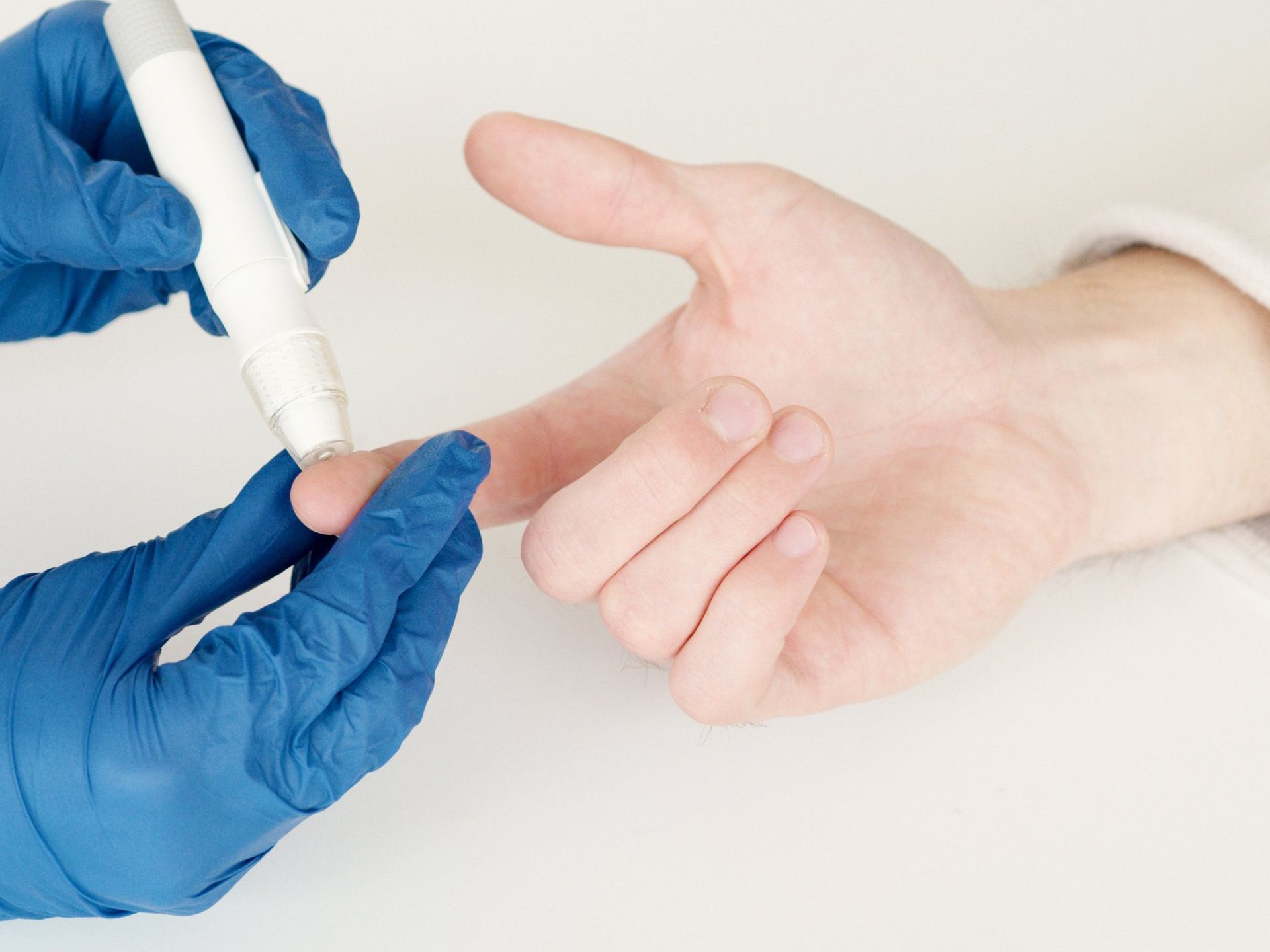 A person wearing blue medical gloves uses a lancet device to prick a finger for a blood glucose test.