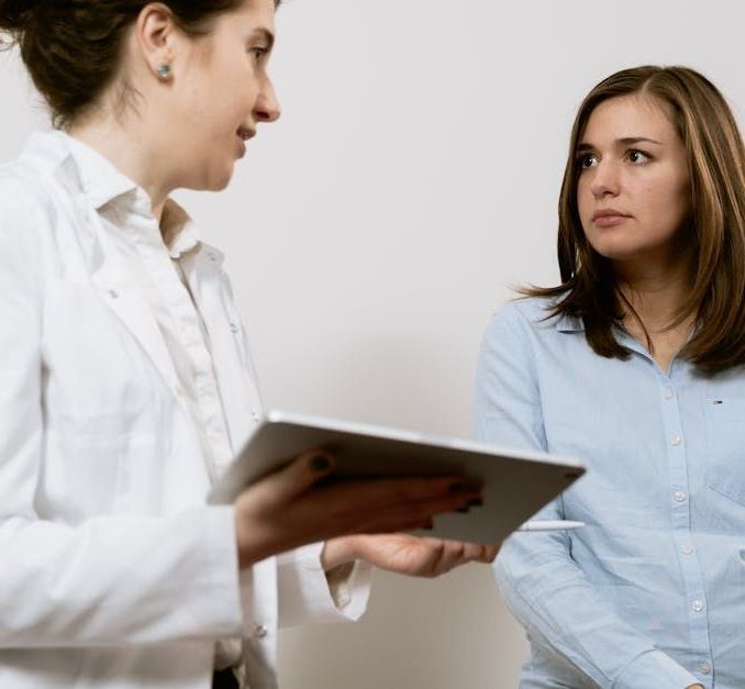 A professional in a white coat holds a tablet while talking to a person in a light blue shirt.