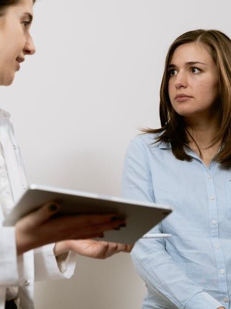 A professional in a white coat holds a tablet while discussing information with a person in a light blue shirt.