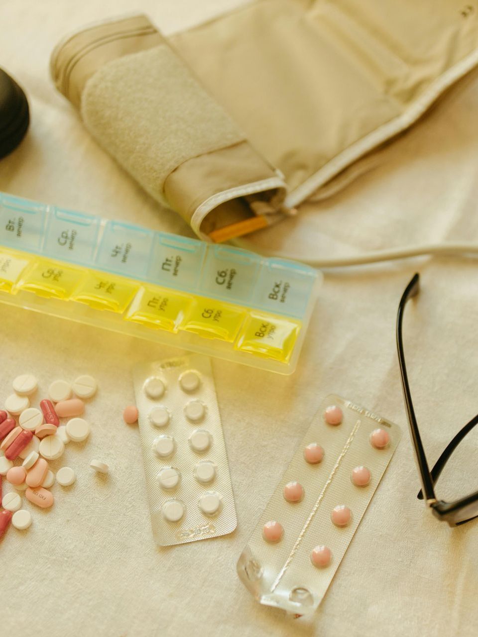 A daily pill organizer, assorted loose medication, two blister packs of tablets, and a blood pressure cuff on a cloth surface.