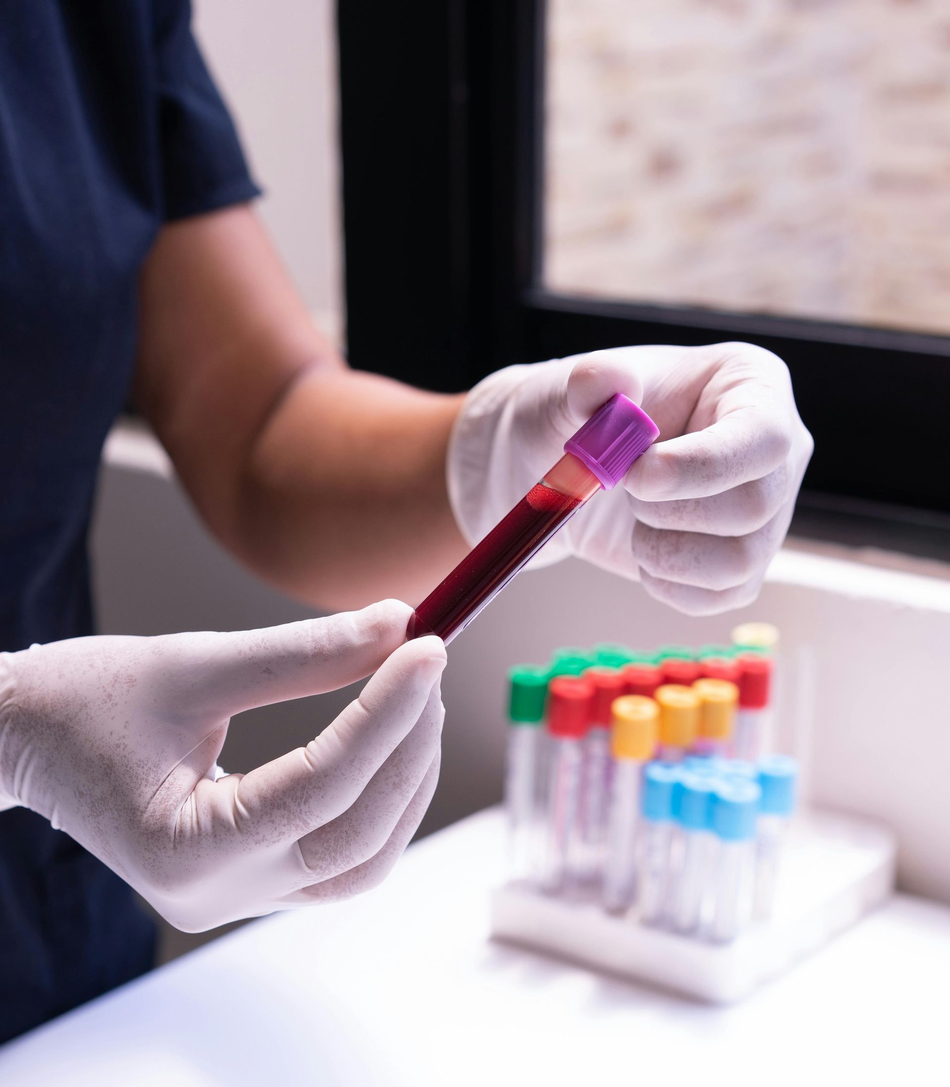 A healthcare professional in gloves holds a purple-topped vial of blood, with a rack of various sample tubes in the back.