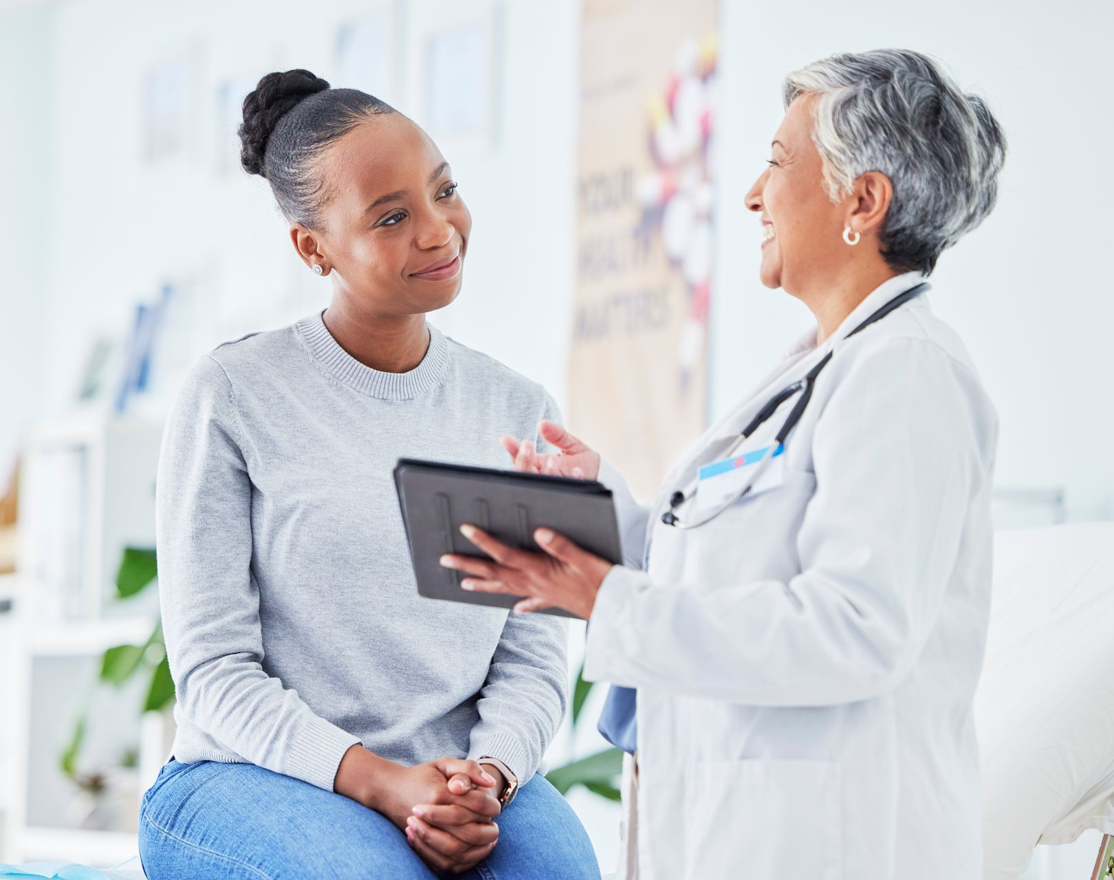A doctor holding a tablet speaks to a patient in an office setting.