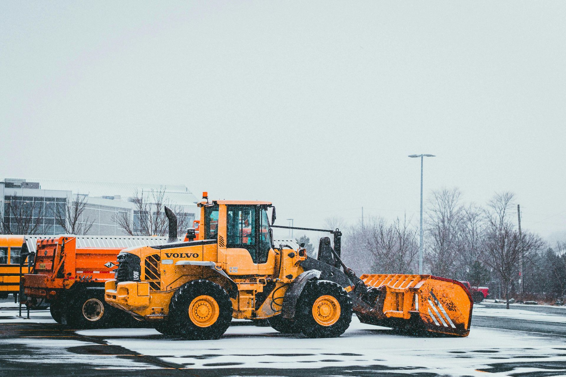 A yellow snow plow is parked in a parking lot next to a school bus.