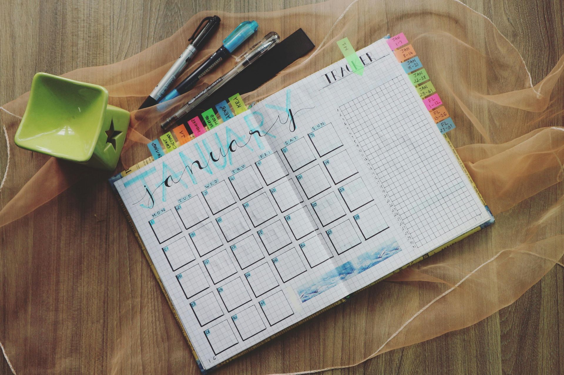 A calendar is sitting on a wooden table next to a cup and pens.