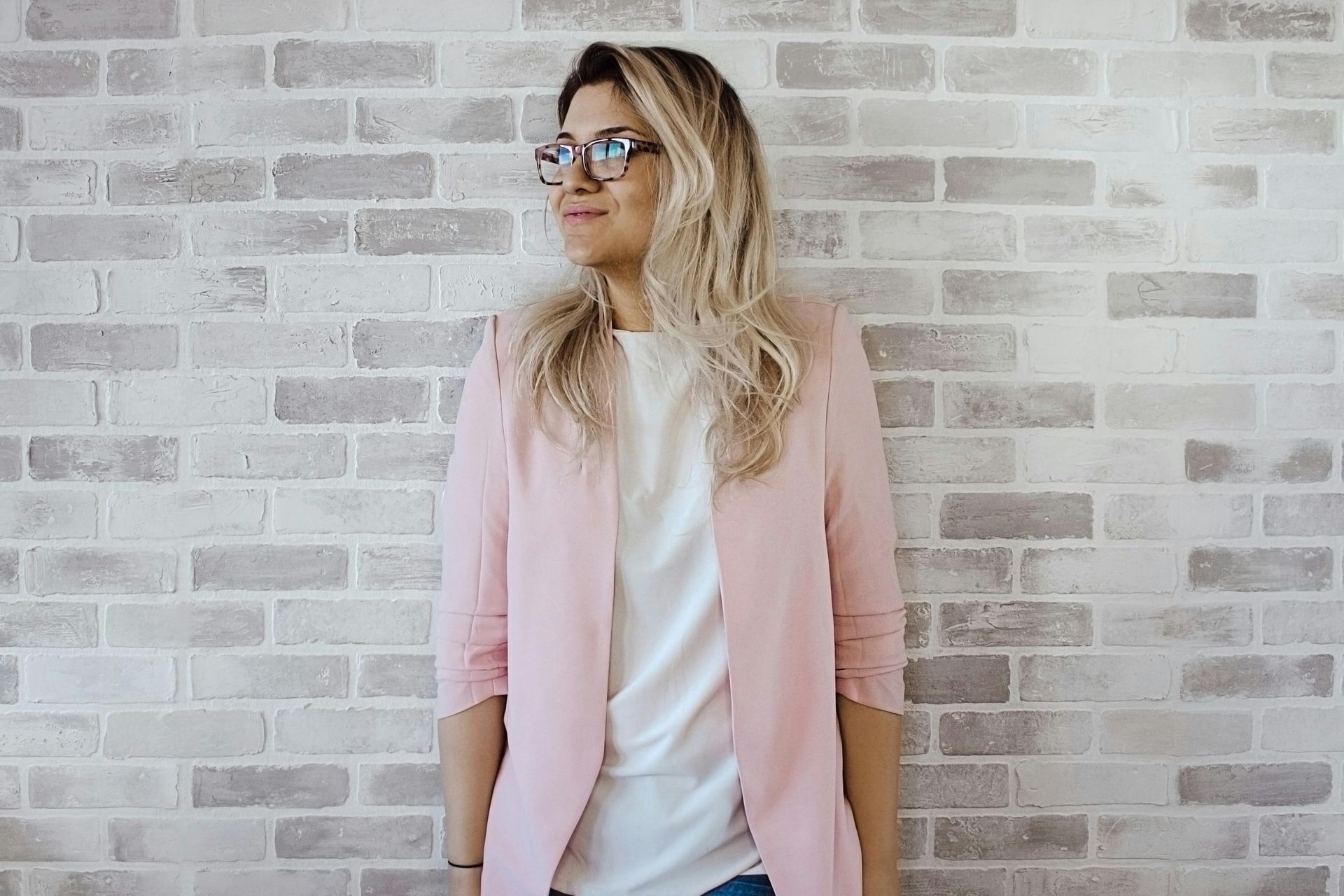 Woman with glasses in a blazer stands against a wall