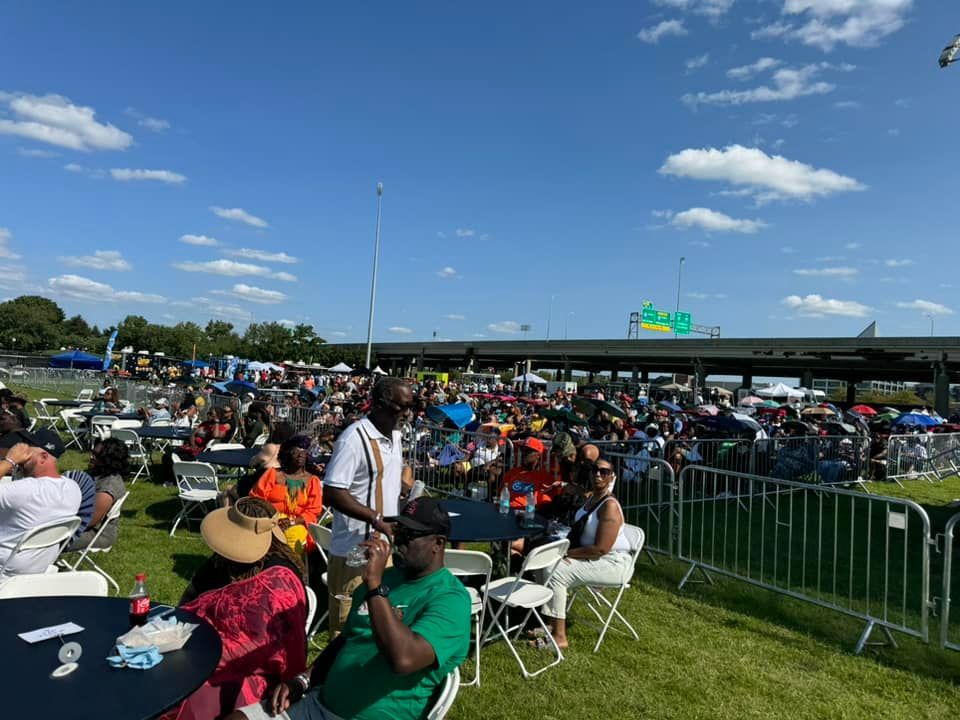 Outdoor festival with a crowd, tables, and a highway overpass in the background under a blue sky.