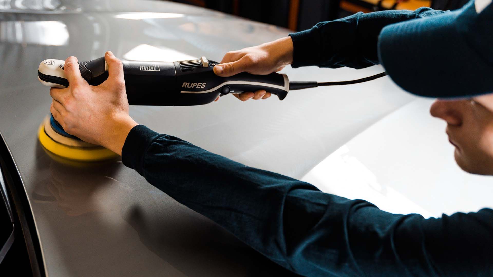 Person using a polisher on a car's surface. The machine is black, and the person wears a hat.