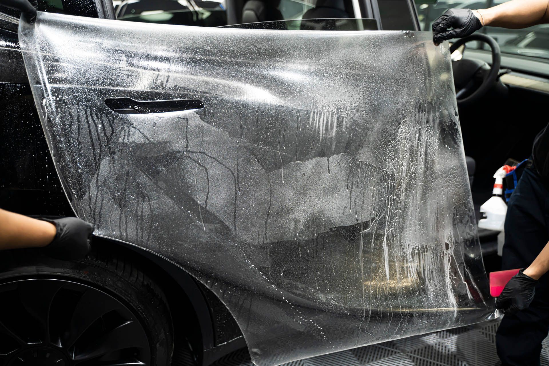 Applying clear protective film to a car door; two people hold up wet, transparent material.