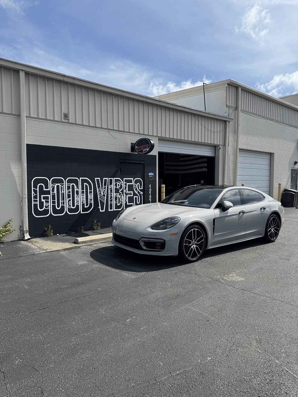Silver sports sedan parked outside an industrial garage with graffiti reading “GOOD VIBES”