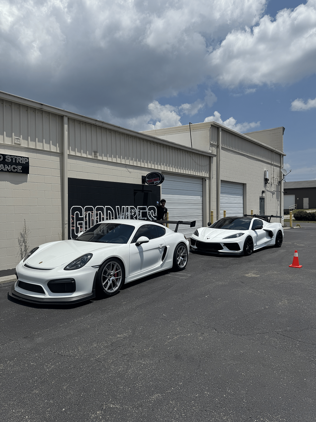 Two white sports cars parked outside a garage with industrial buildings and cloudy sky behind them