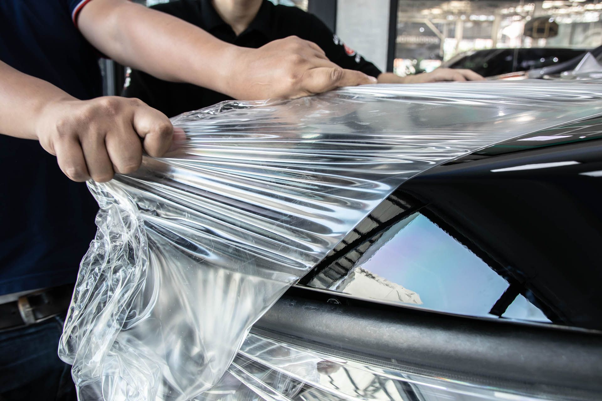 Two people applying clear plastic wrap to a black car, likely for protection.