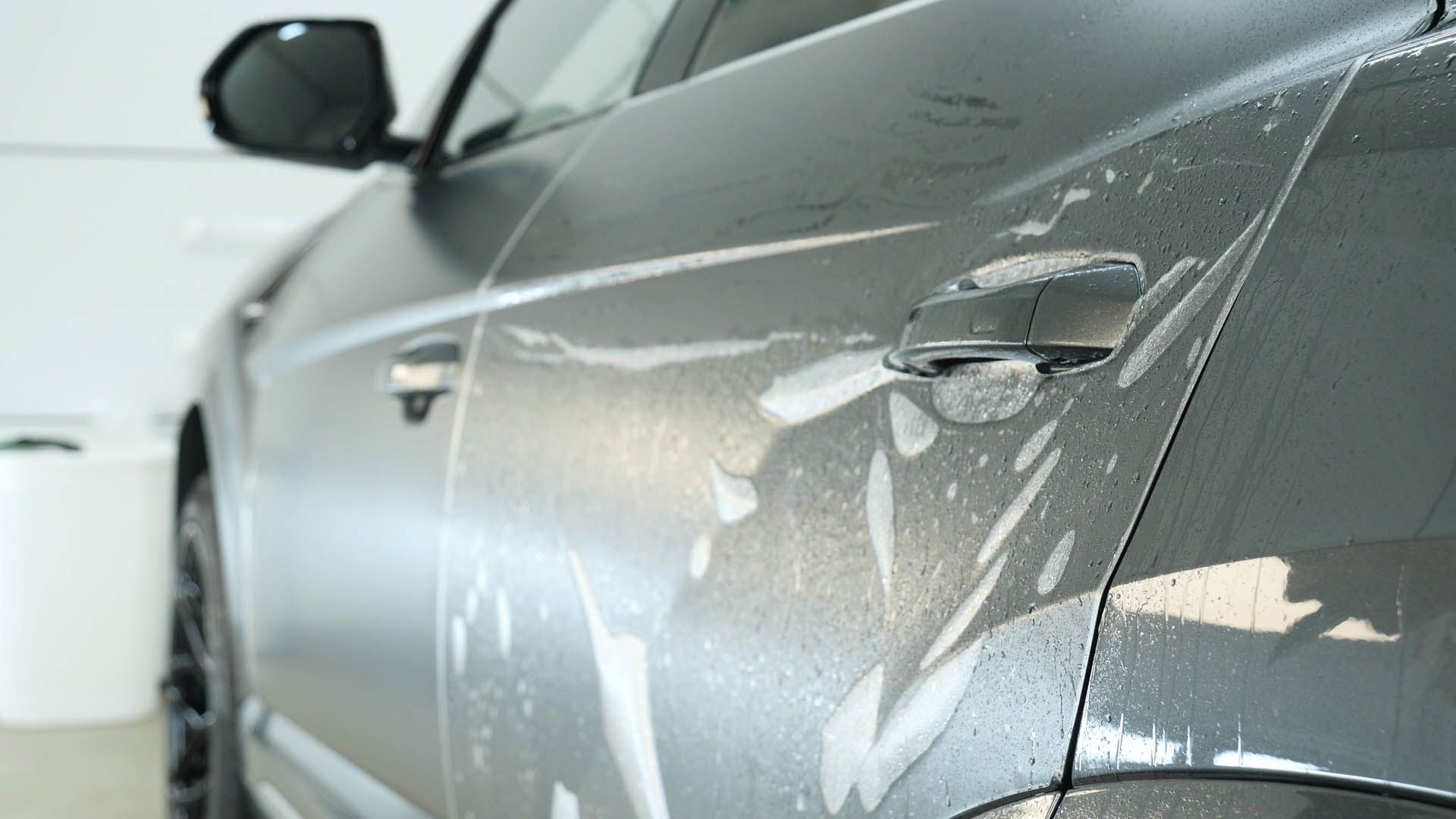 Gray car door covered in soapy foam, being washed indoors.