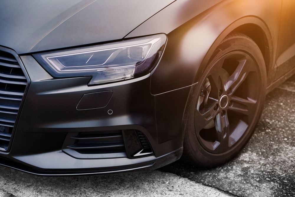 Black car, close-up of front. Headlight detail, black rims, parked on asphalt with sunlight.