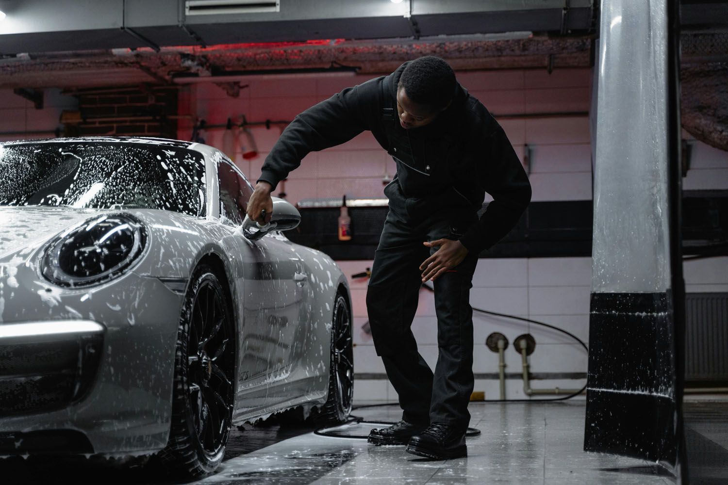 Person washing a silver car with black wheels, covered in soap suds, in a garage.