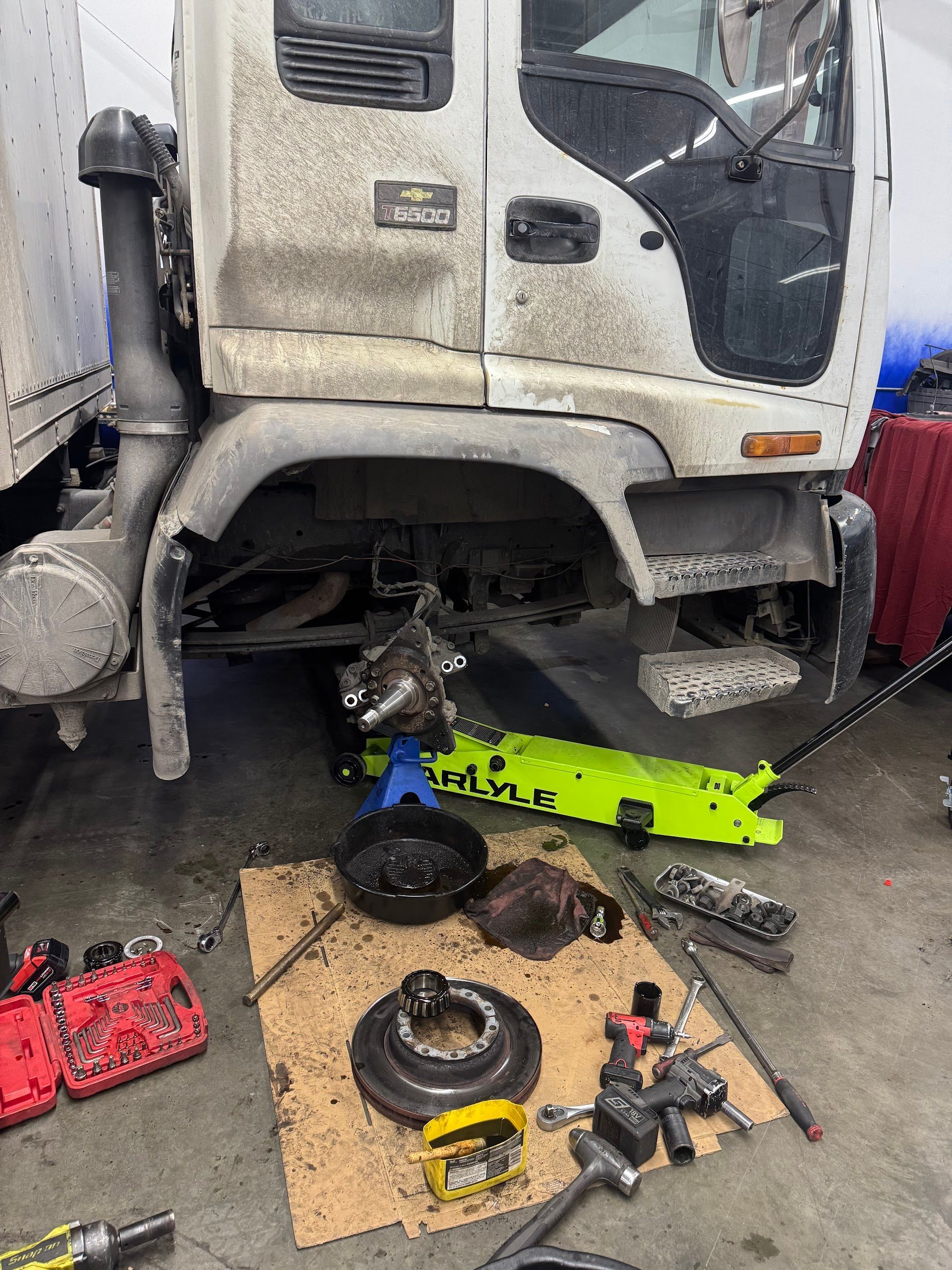 A white Chevy truck under repair in a garage with its wheel removed, a bright green floor jack, and tools on cardboard. | G Tech Repair