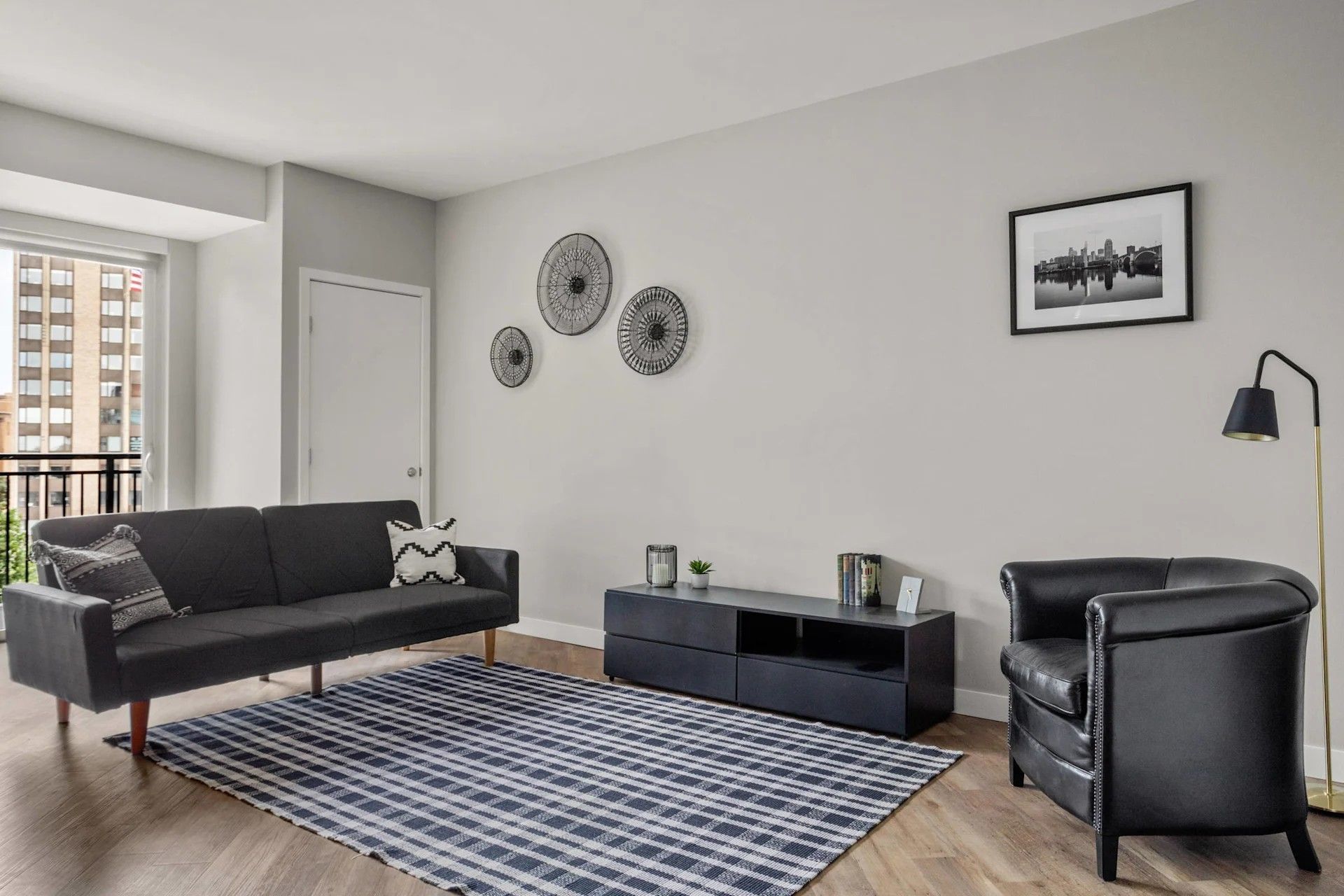 A modern living room with a black sofa, a patterned rug, a dark media console, a leather armchair, and wall decor.
