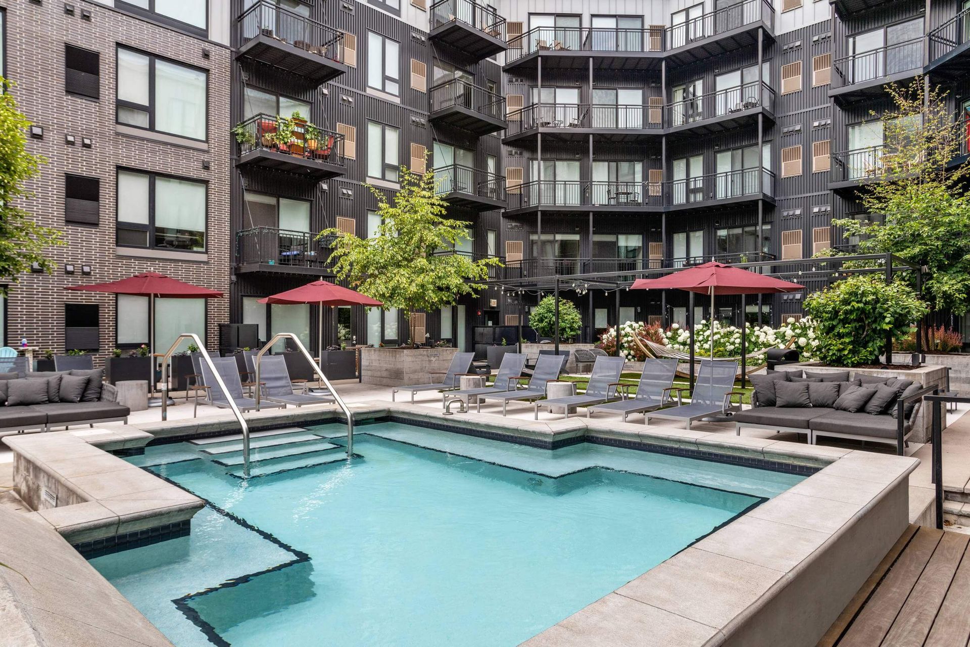 A swimming pool courtyard at a modern multi-story apartment building with patio furniture and red umbrellas.