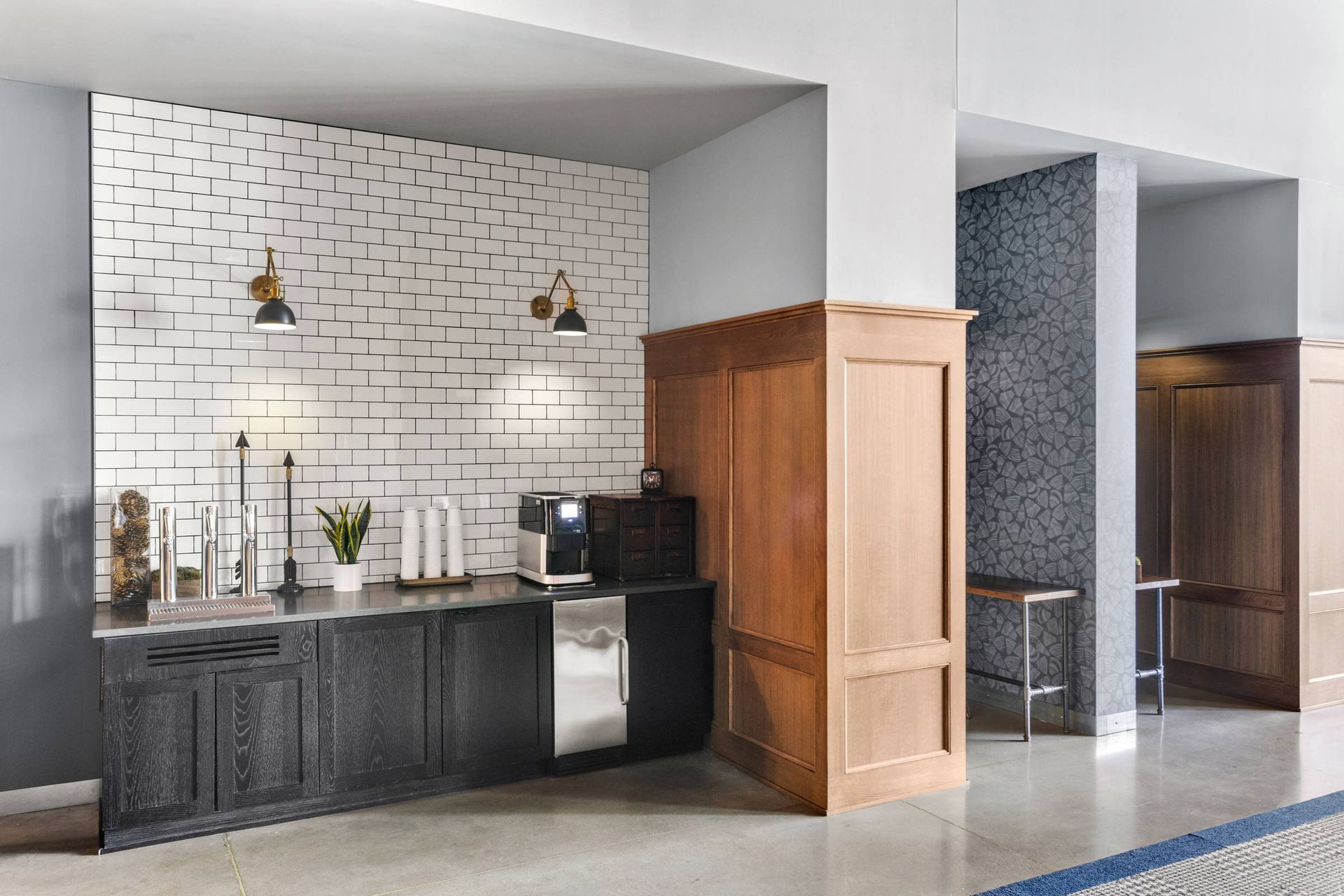 A modern coffee station with a patterned tile wall, black cabinets, and two wall sconces in a hallway with wooden accents.