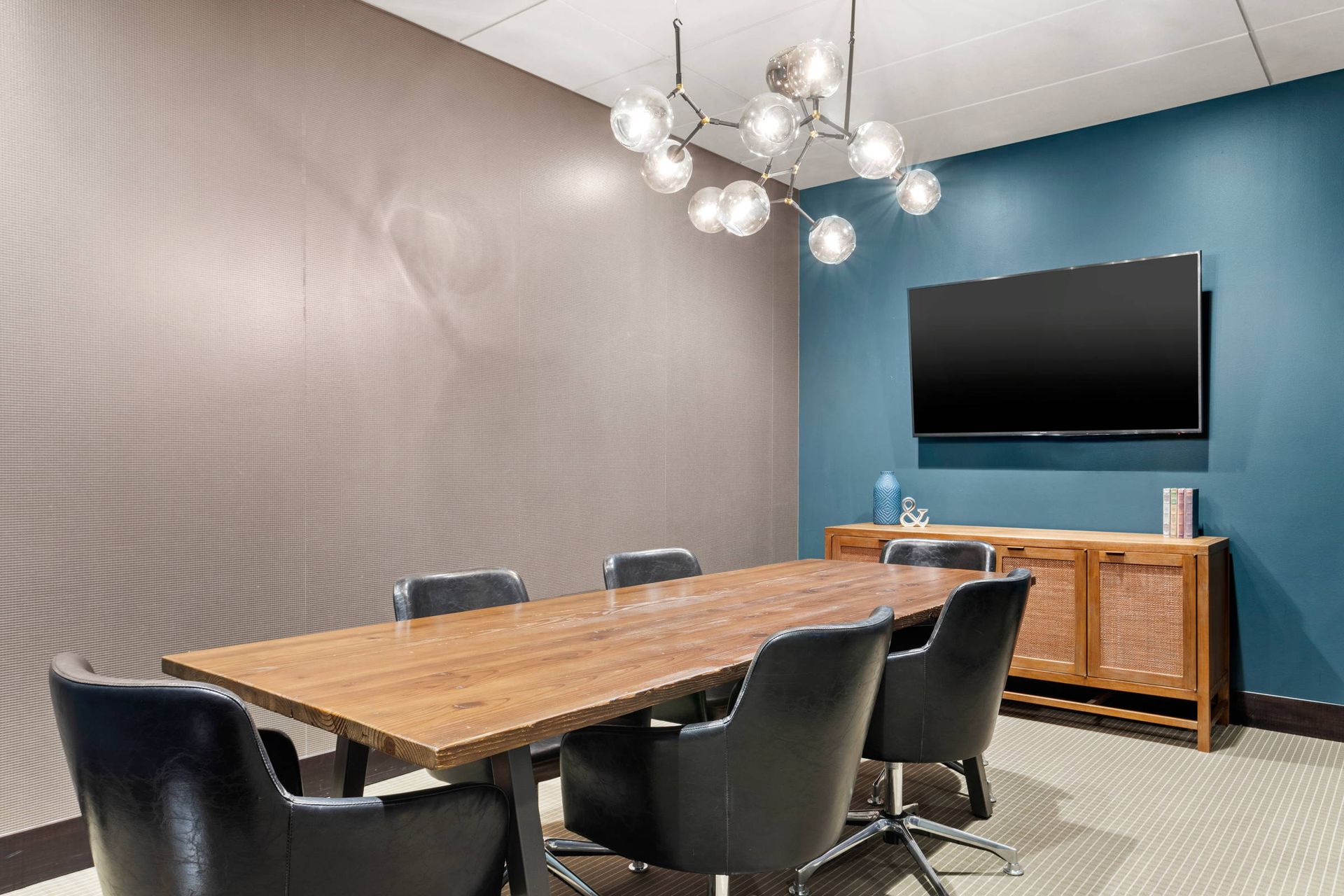 Modern conference room with a wooden table, black chairs, a teal accent wall with a TV, and a globe-style chandelier.