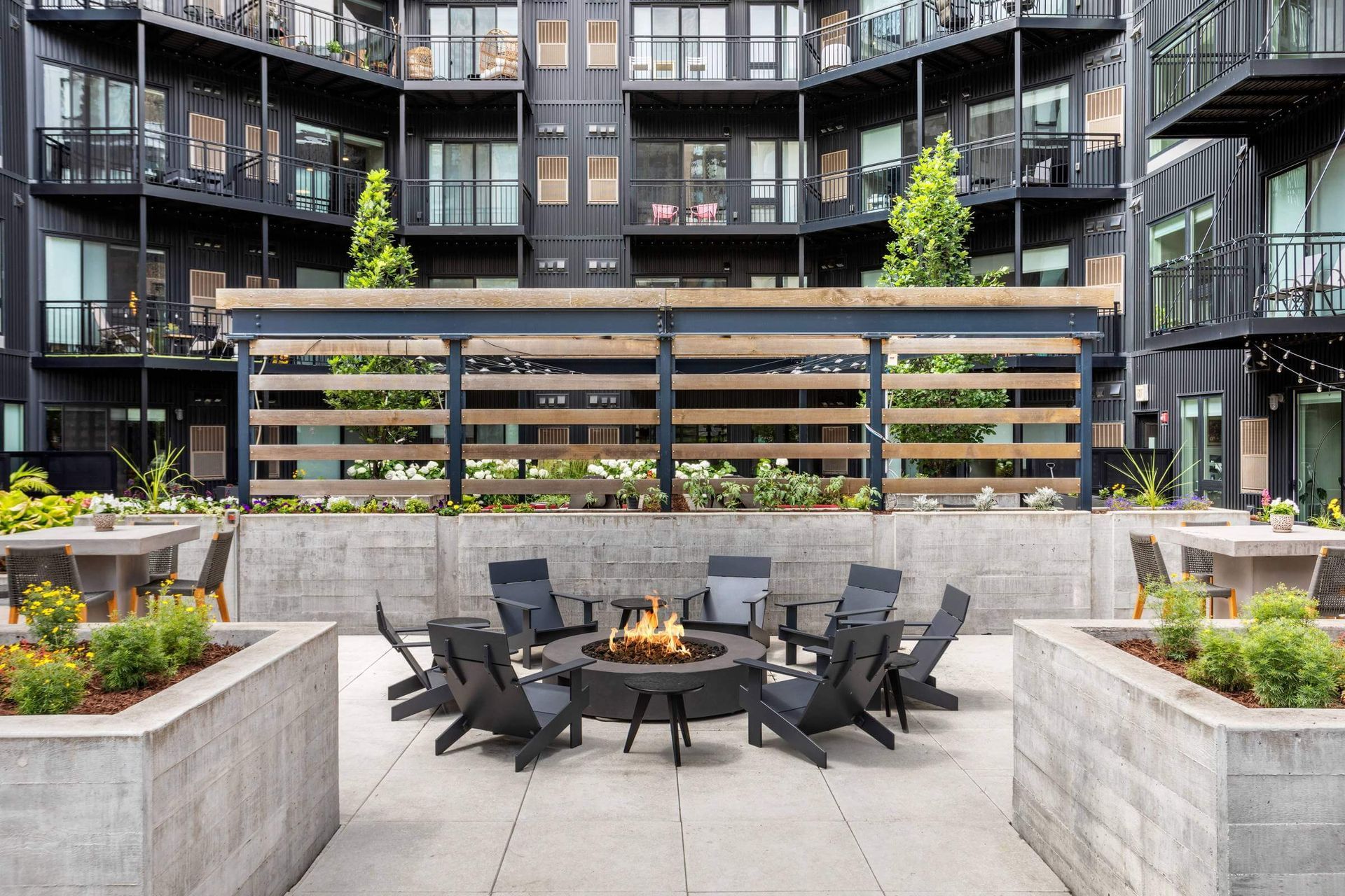 A courtyard with a fire pit surrounded by dark chairs, flanked by concrete planters and a modern apartment building.