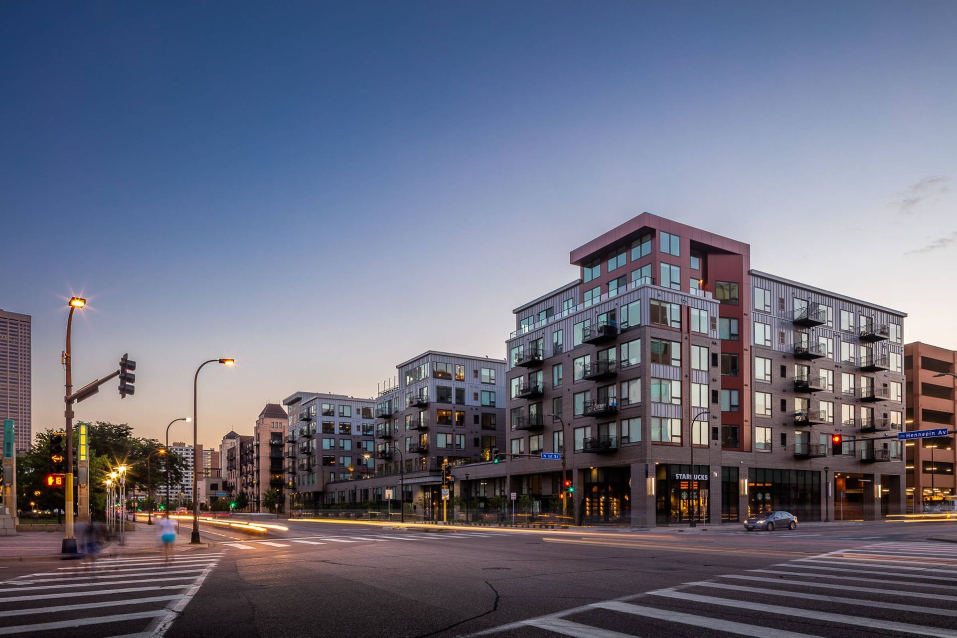 A modern multi-story apartment building with balconies and ground-floor retail at a street corner during twilight.