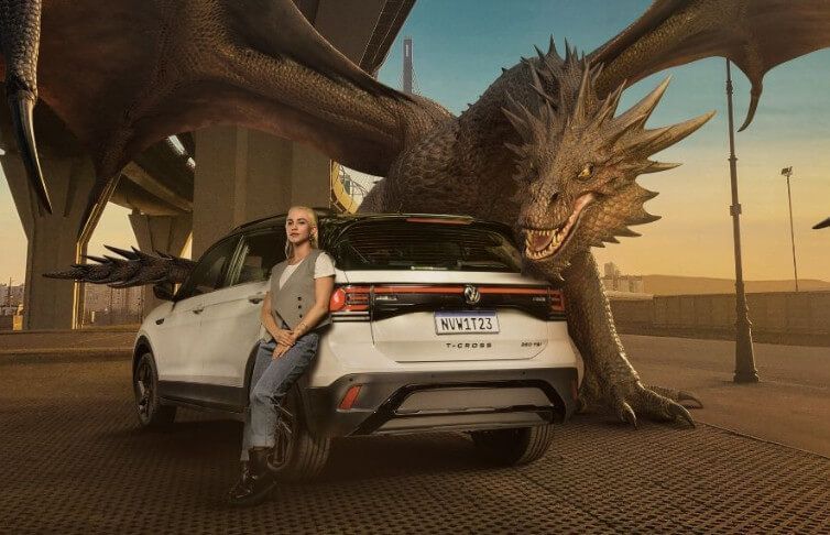 Homem encostado em um SUV branco ao lado de uma escultura gigante de dragão ao pôr do sol