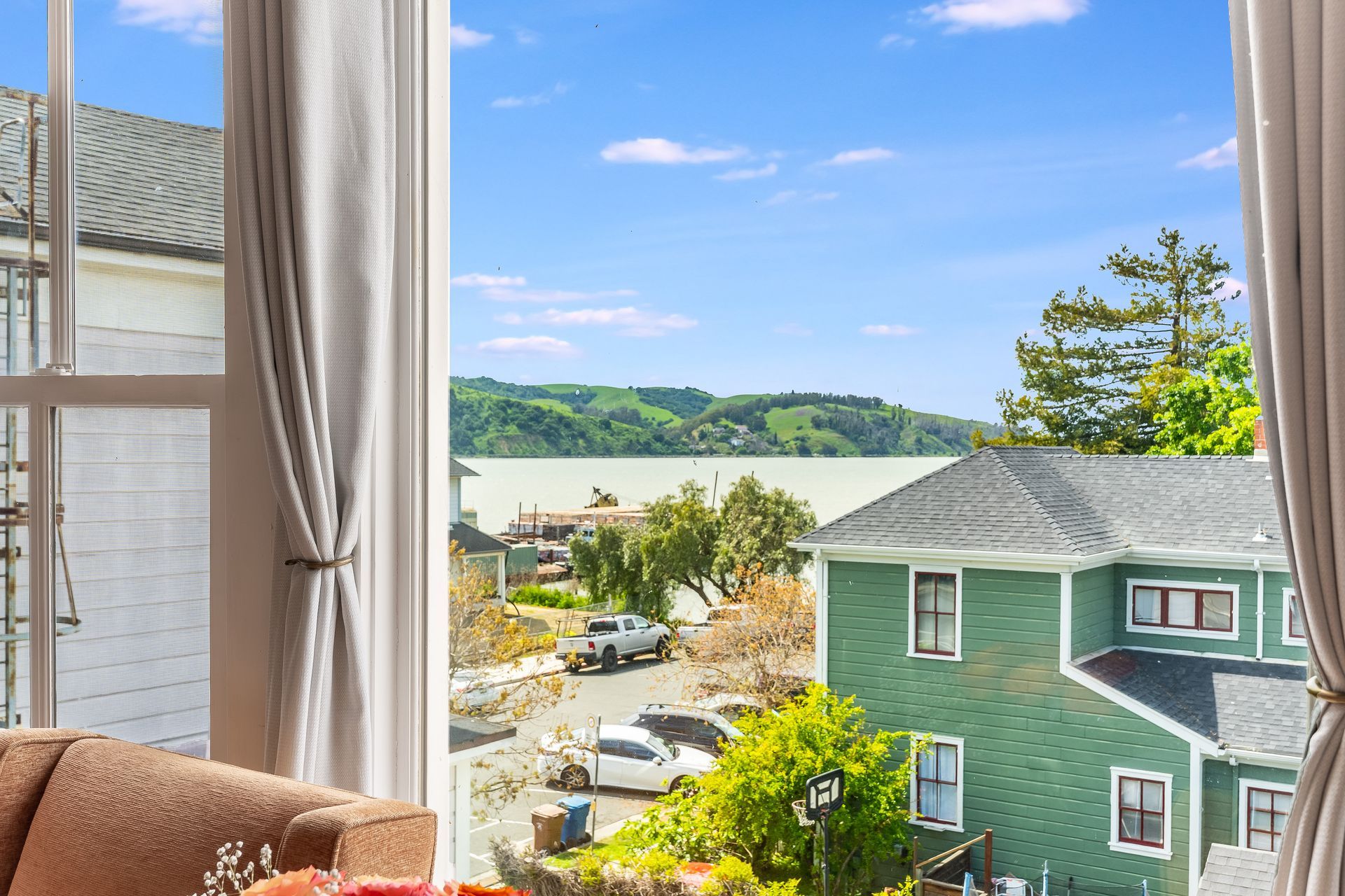 View through a window: ocean and houses under blue sky, with curtains framing the scene.